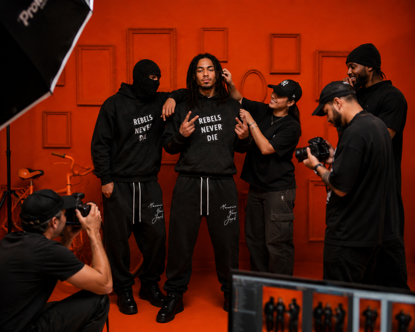 MXNY streetwear photoshoot behind the scenes: five people in black Rebels Never Die hoodies posing and shooting photos against a red wall.
