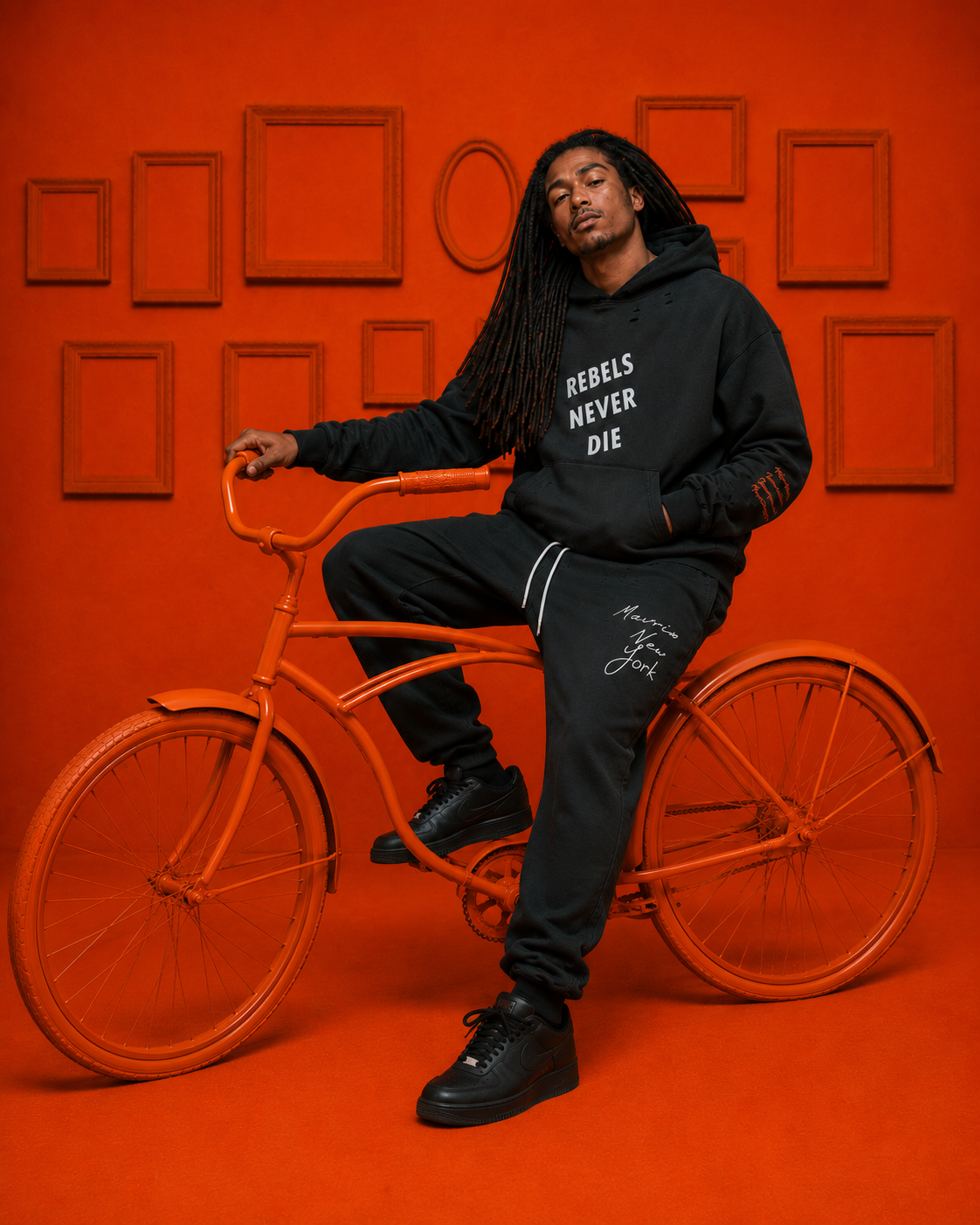 A man with long dreadlocks sitting on an orange bicycle in front of an orange wall with empty picture frames. He is wearing a black hoodie with the words 'REBELS NEVER DIE' and black pants with 'Morris New York' written on them.