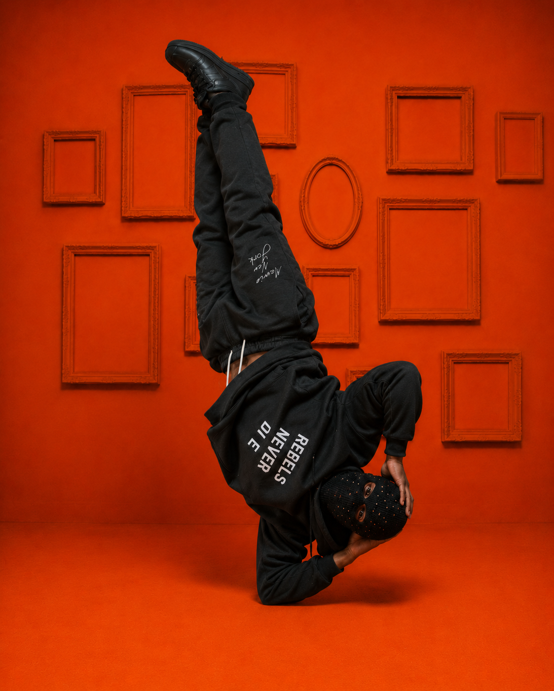 MXNY model performing a headstand in an all-black Rebels Never Die hoodie and sweatpants against an orange backdrop, showcasing bold Bronx streetwear.