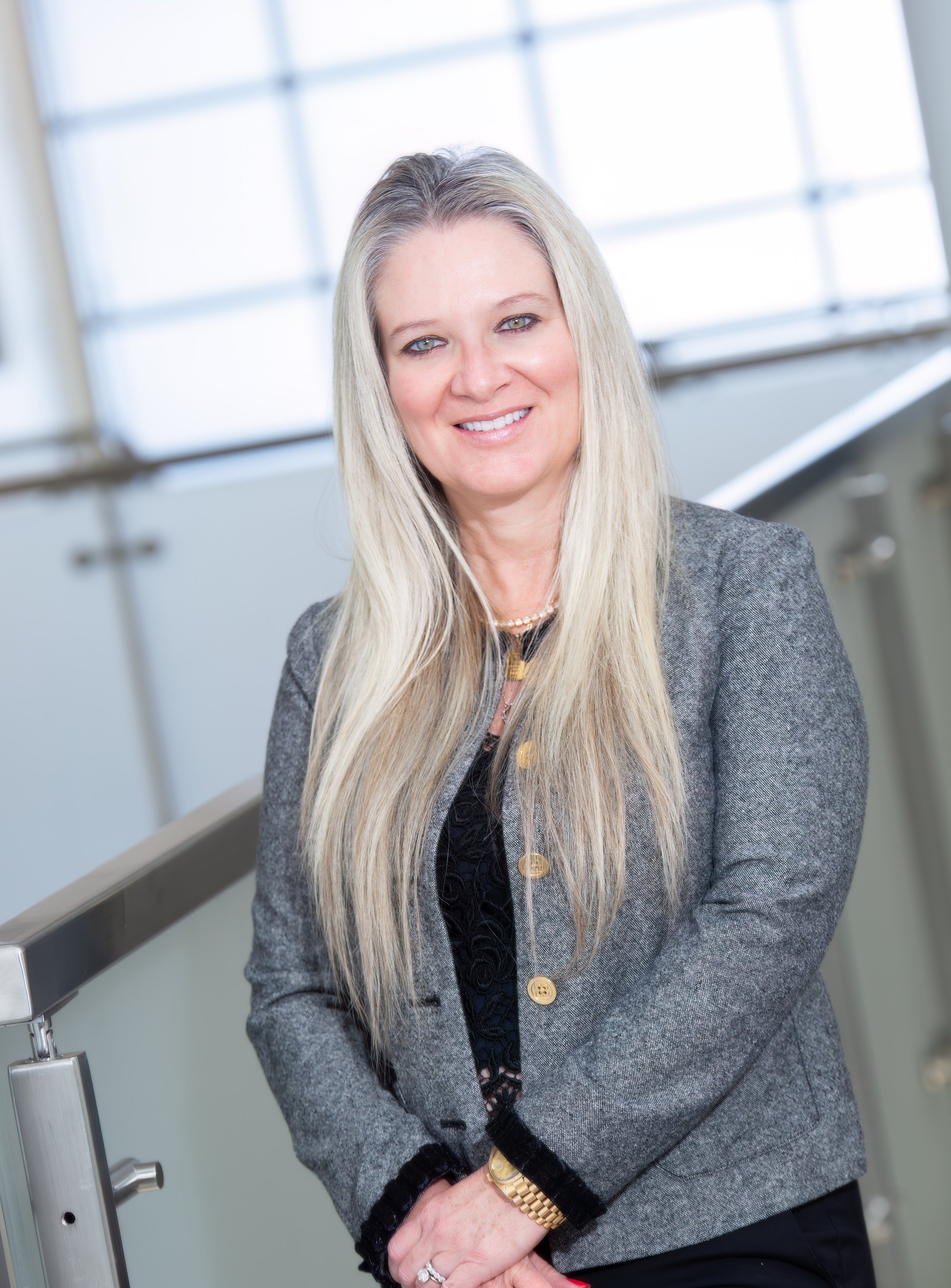 A professional woman with long blonde hair, wearing a grey blazer, black top, and gold jewelry, standing near a railing in a bright room with large windows.