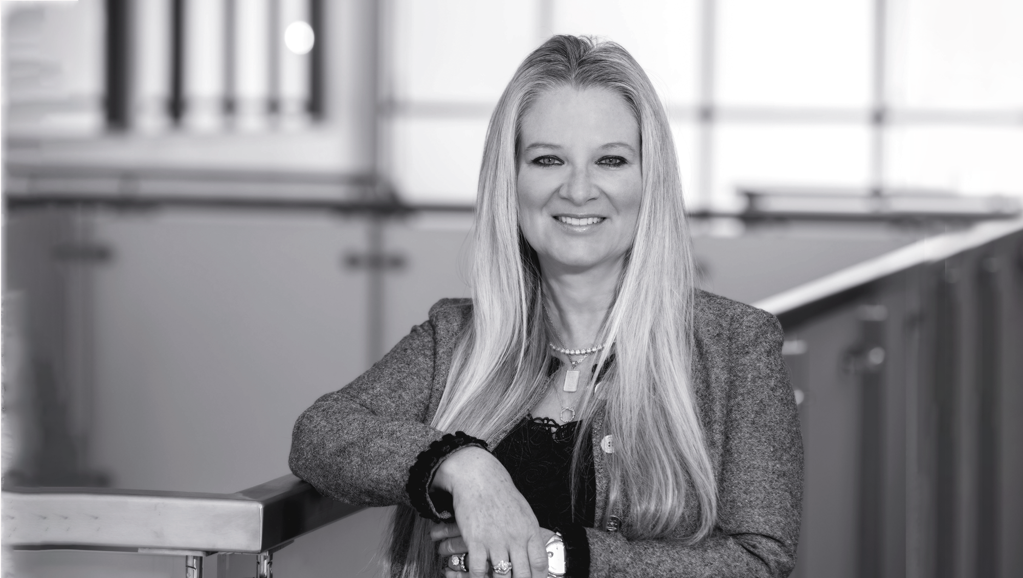 Black and white portrait of a woman with long, straight hair, smiling, sitting at a table inside an office or modern building.