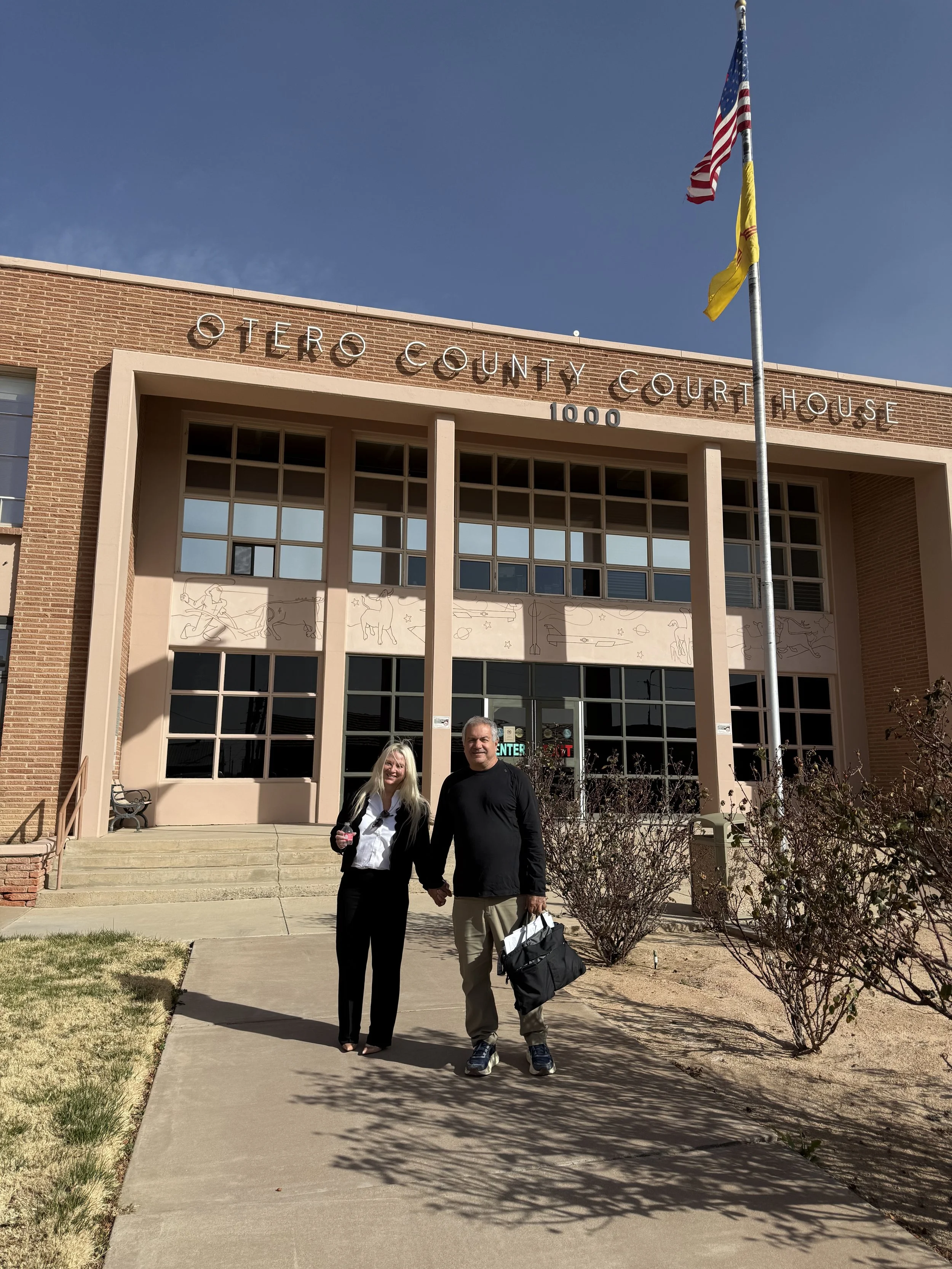 A man and woman holding hands standing in front of Cerro County Courthouse, with an American flag and a yellow flag flying on a flagpole nearby.