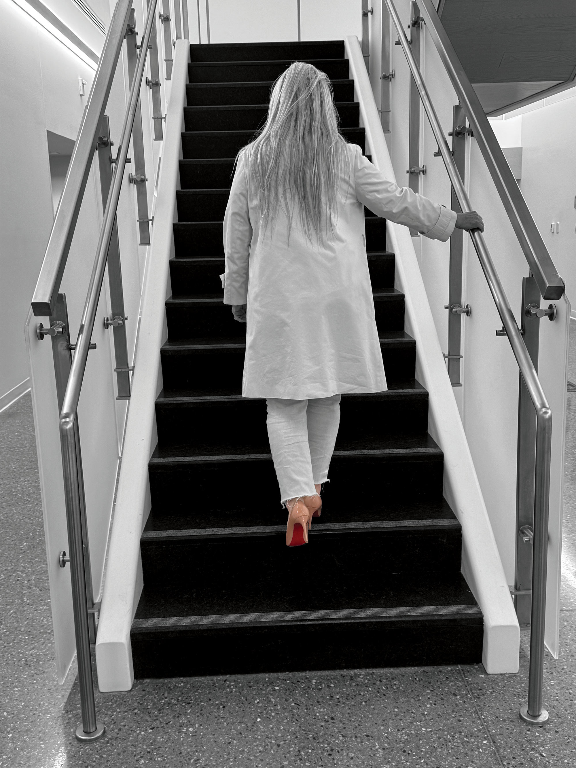 Woman with long hair in a white coat and high heels walking up stairs at office building.