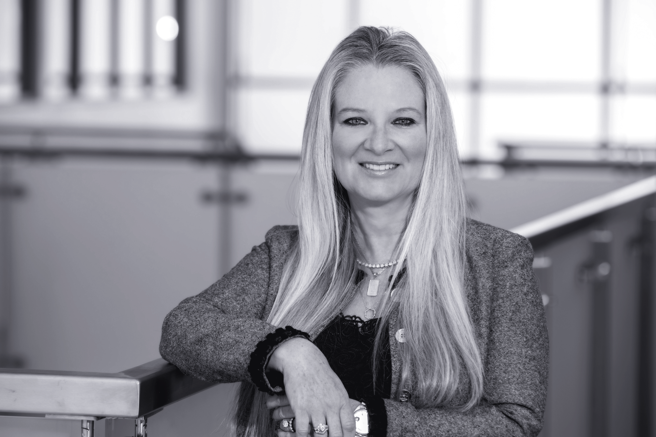 Black and white photo of a woman with long hair smiling at the camera, sitting at a desk with a metal edge in an office setting.