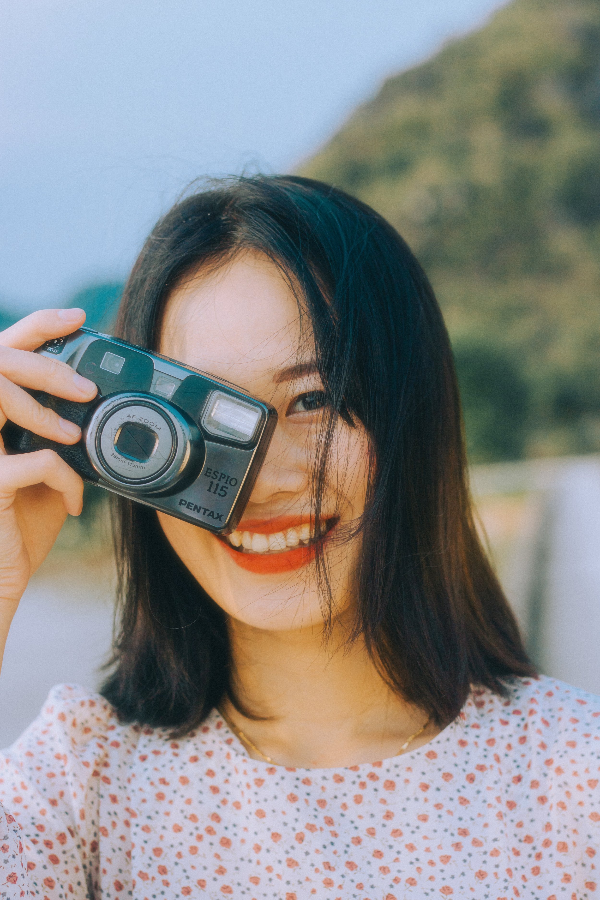 A woman with black hair smiling while holding a vintage camera in front of her face outdoors during daytime.