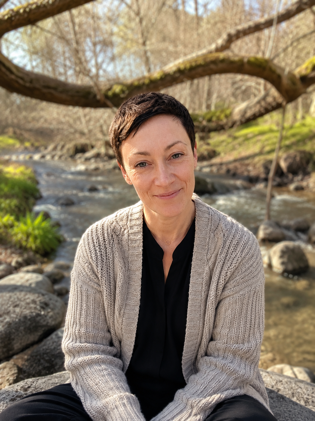 A woman with short dark hair sits on rocks by a creek, smiling at the camera, with a large tree arching overhead in a natural outdoor setting.