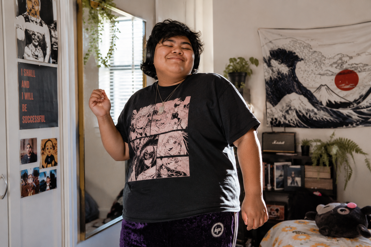 A person with curly black hair wearing headphones and a black graphic t-shirt with anime characters, smiling and dancing in a room decorated with posters, art, and plants.