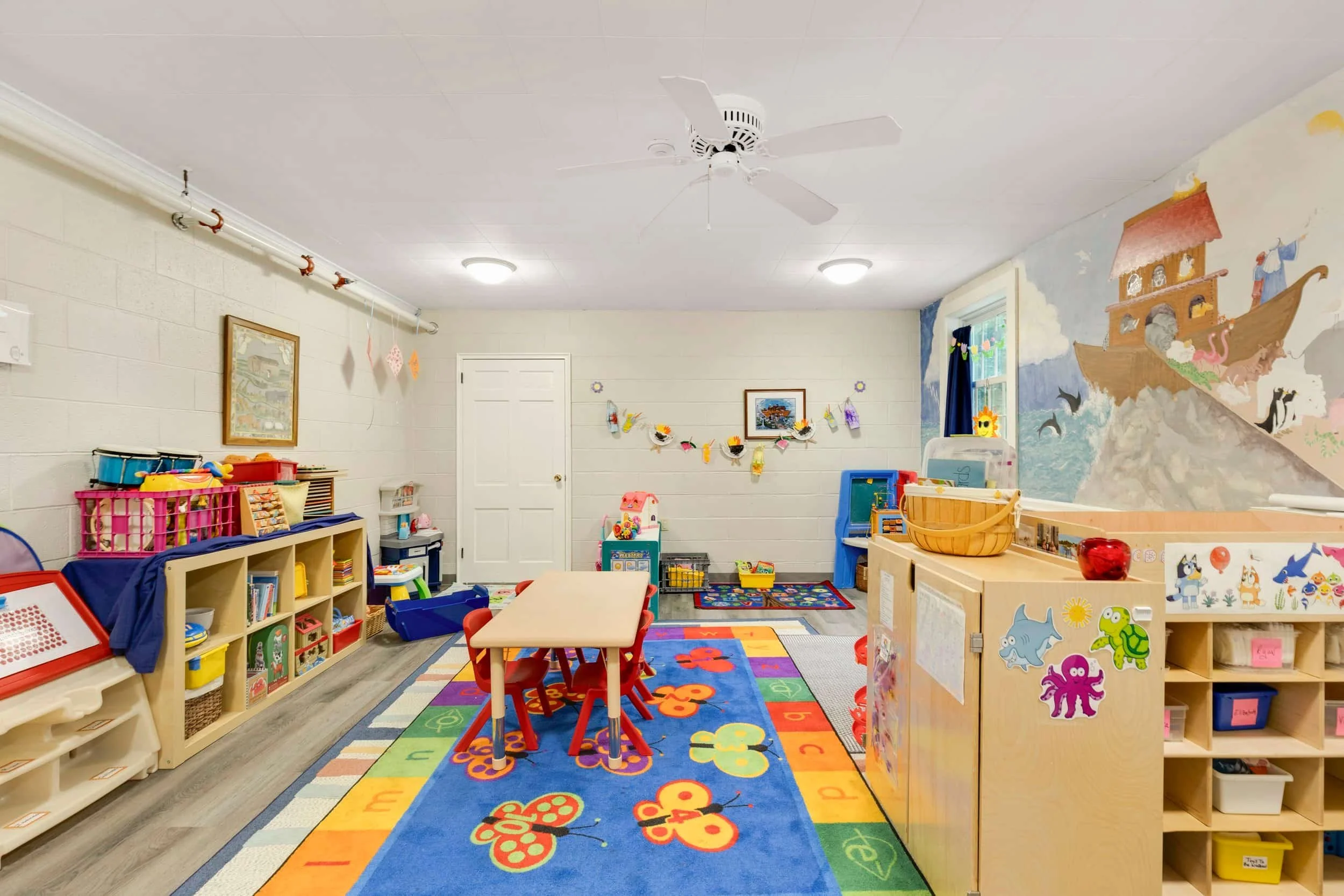 A colorful playroom with a rainbow-colored alphabet rug, small tables and chairs, shelves filled with toys and books, a wall mural of a ship and animals, and decorations hanging from the ceiling.