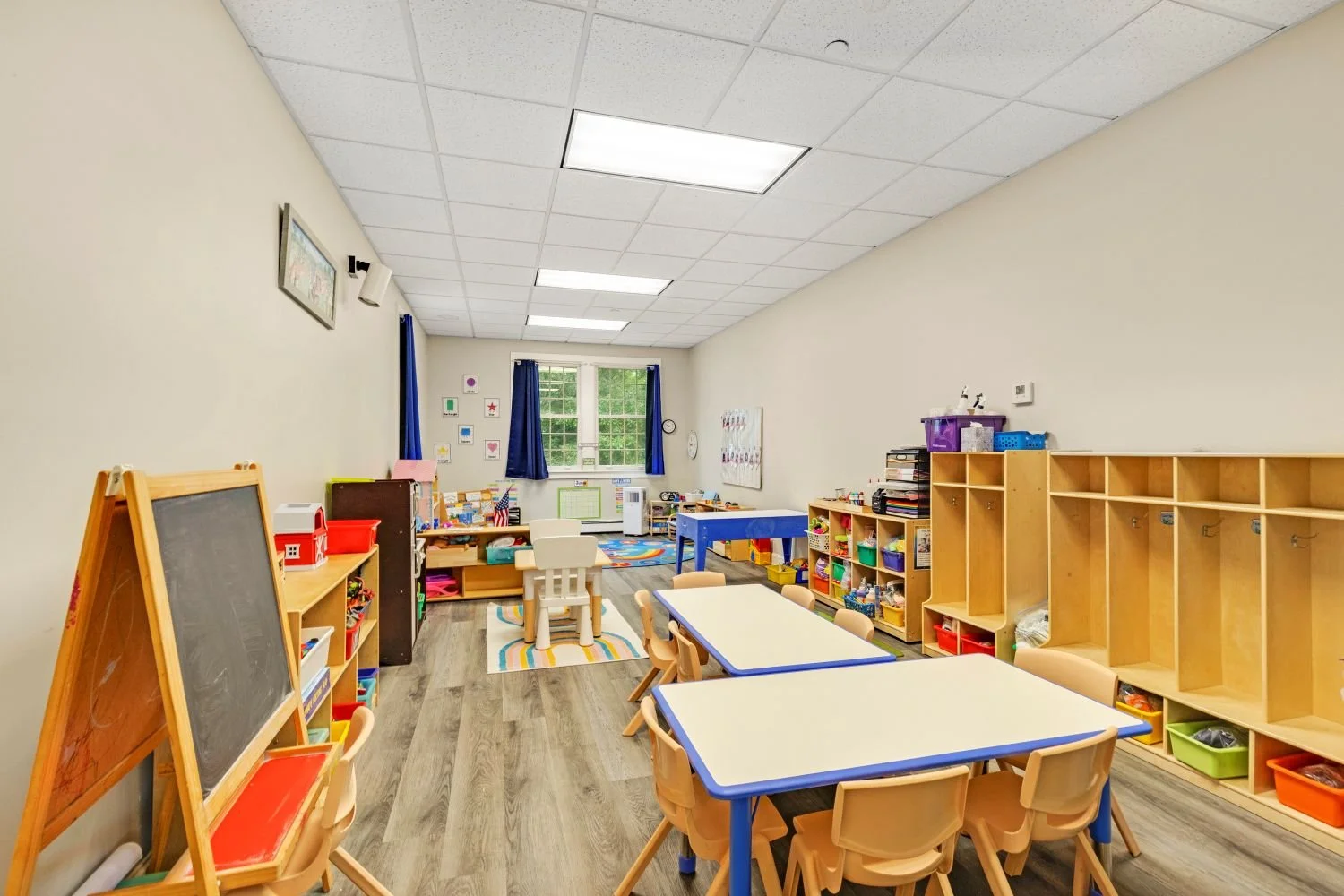 A bright preschool classroom with tables and chairs, educational toys, and artwork on the walls.