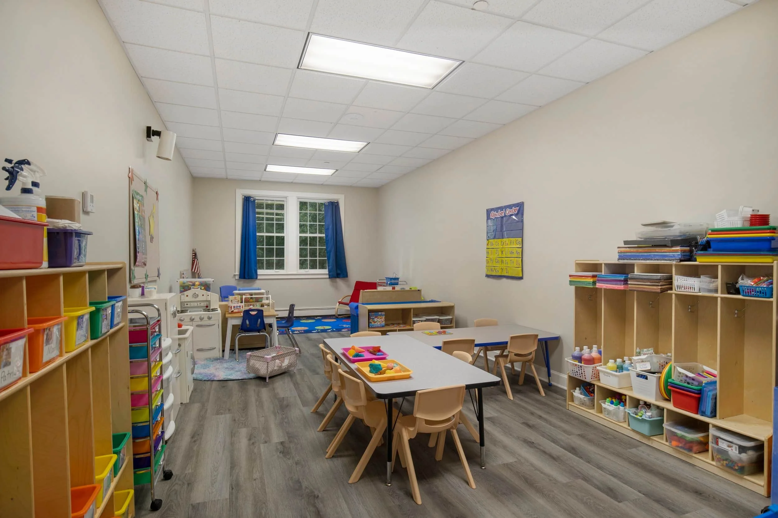 A classroom with bookshelves, tables, chairs, and educational materials, illuminated by fluorescent ceiling lights and a window with blue curtains.