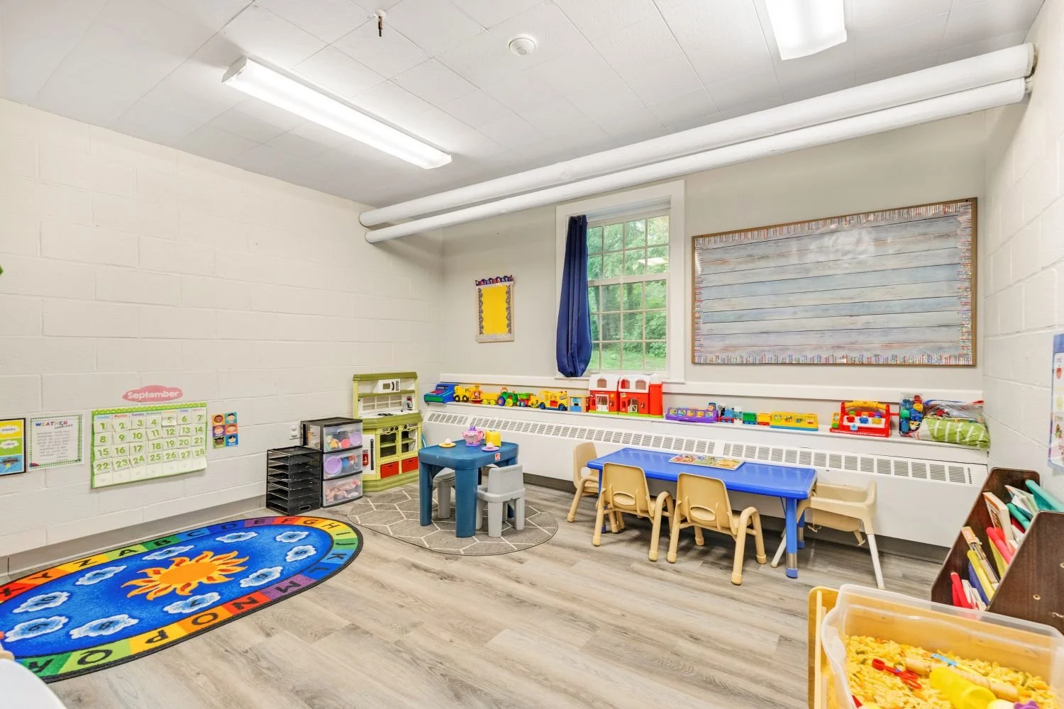 A colorful preschool classroom with small tables, chairs, toys, and educational materials, a rainbow-colored rug with a sun design, and a window with a blue curtain.