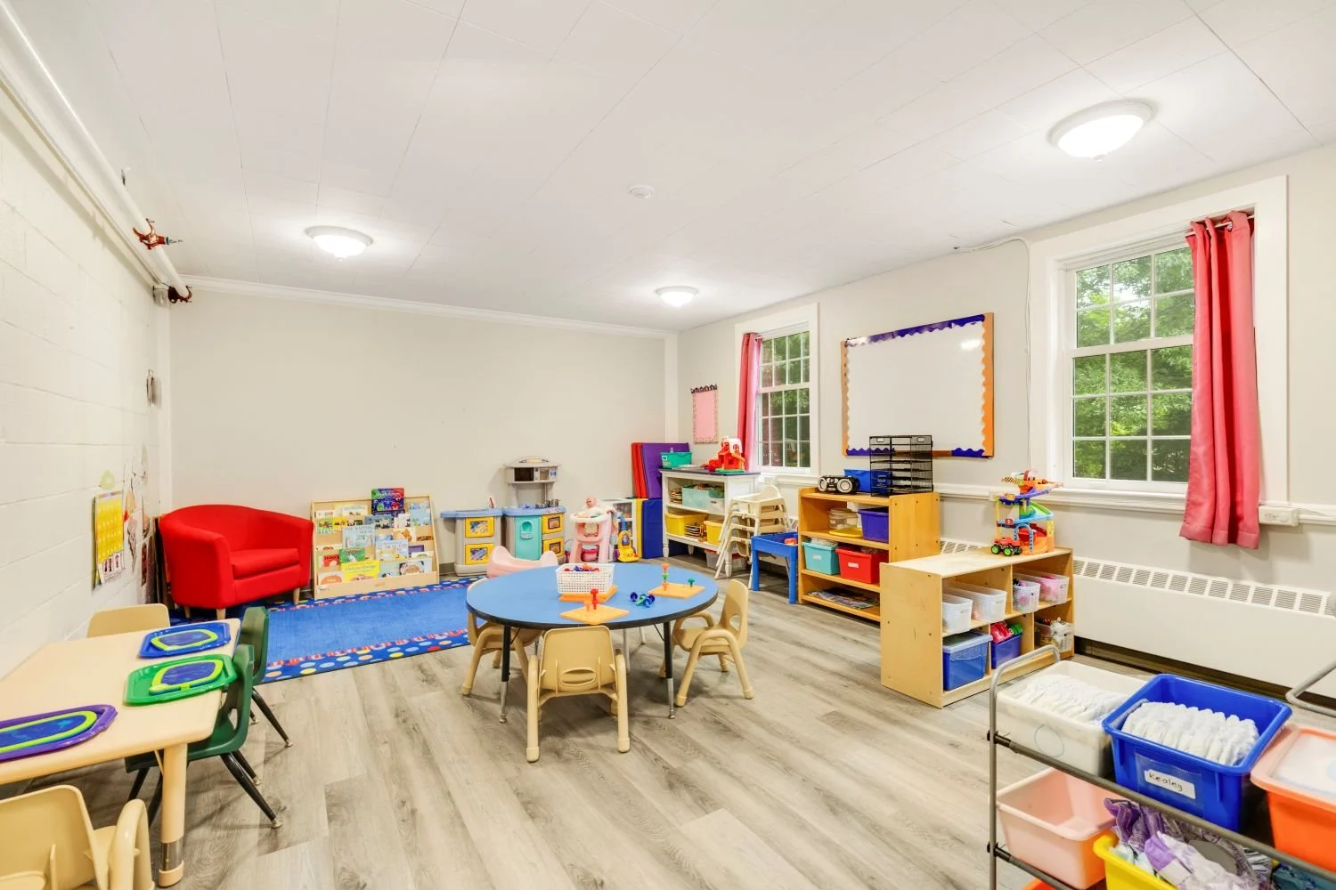 A brightly lit preschool classroom with colorful furniture, toys, and learning materials, including a round table with chairs, a red armchair, storage shelves, and windows with pink curtains.