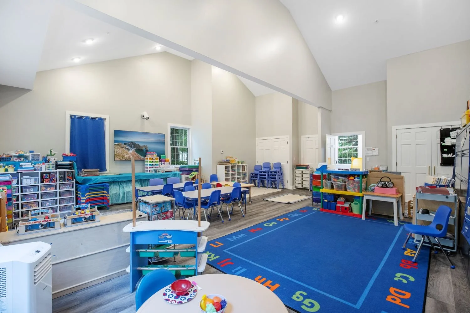 Bright classroom with blue carpet featuring colorful alphabet border, small tables and chairs, and various educational materials and toys, illuminated by natural light from windows.