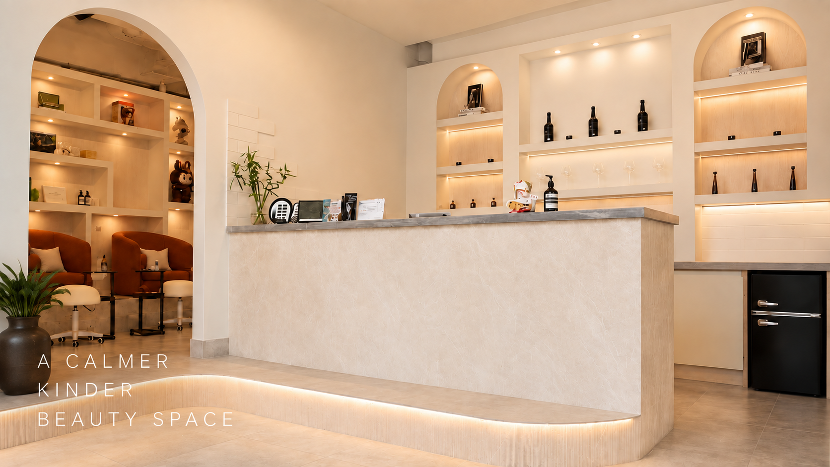 A minimalist beauty space with a reception desk, warm lighting, and decorative shelving featuring bottles and art pieces, along with seating area with brown chairs and a potted plant.
