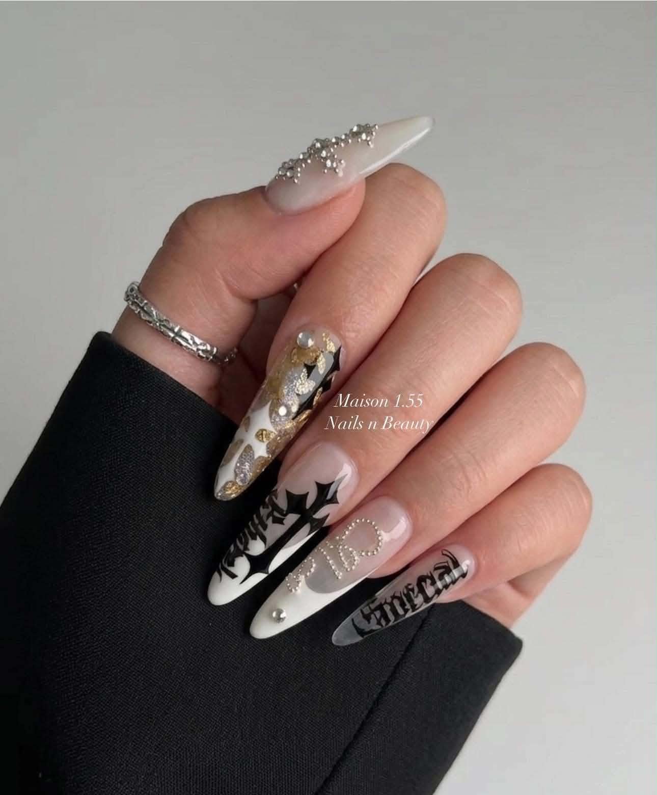 Close-up of a person's hand with long, ornate nails featuring various designs including pearls, black and white patterns, and gold and silver accents. The person is wearing a silver ring and a black sleeve.