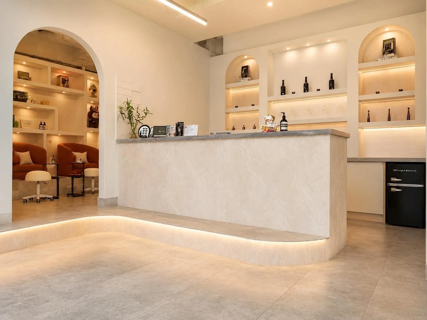 Modern hotel reception area with a curved counter, built-in shelving with bottles and decor, seating area with armchairs and tables, and minimalistic design with soft lighting.
