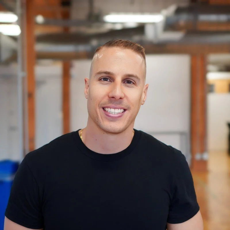 A young man with short light brown hair, smiling, wearing a black T-shirt, indoors in an industrial-style space with exposed beams and shelves in the background.