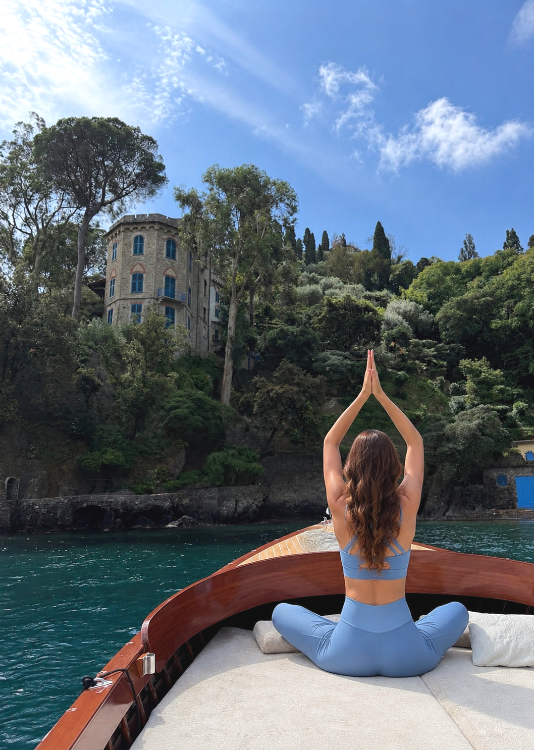 A woman practicing yoga on a boat with a scenic hillside, lush greenery, a castle-like house, and a partly cloudy blue sky in the background.