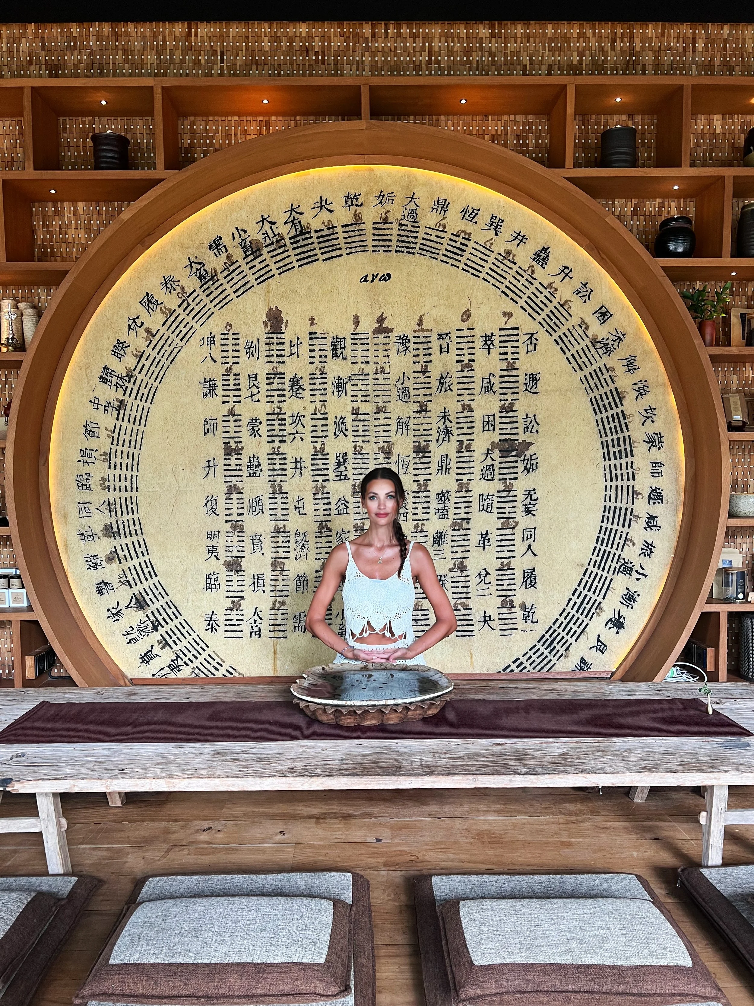 A woman in a white lace top is standing in front of a large decorative wall with Chinese characters and symbols, holding her hands together in a prayer or meditative pose, in a room with wooden furniture and shelves.
