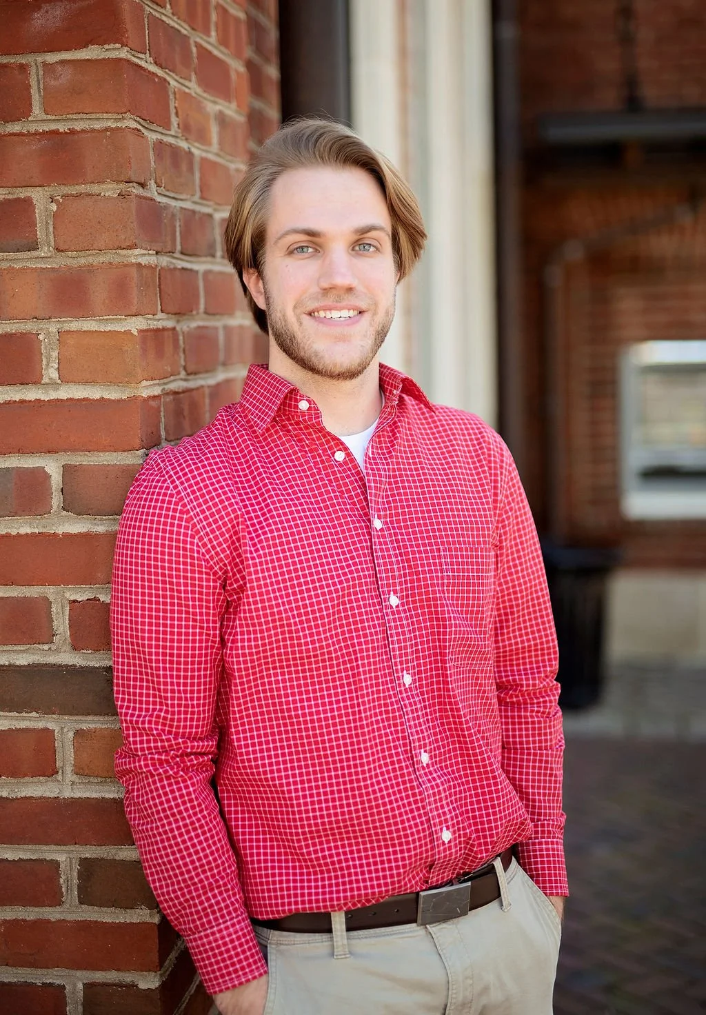A young man with light skin and brown hair, wearing a red checkered shirt and beige pants, standing outdoors against a brick wall, smiling at the camera.