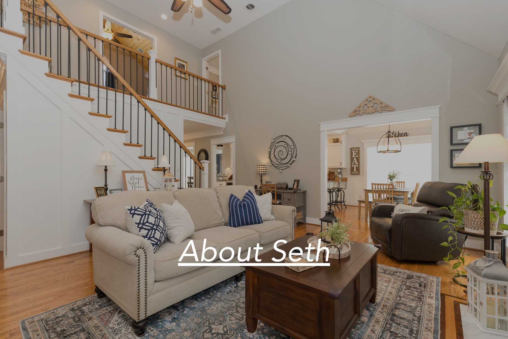 Living room with beige sofa, dark leather chair, wooden coffee table, and decorative pillows; staircase with wooden handrail and black balusters; dining area visible through doorway; various wall art and lamps; hardwood floors and natural light.