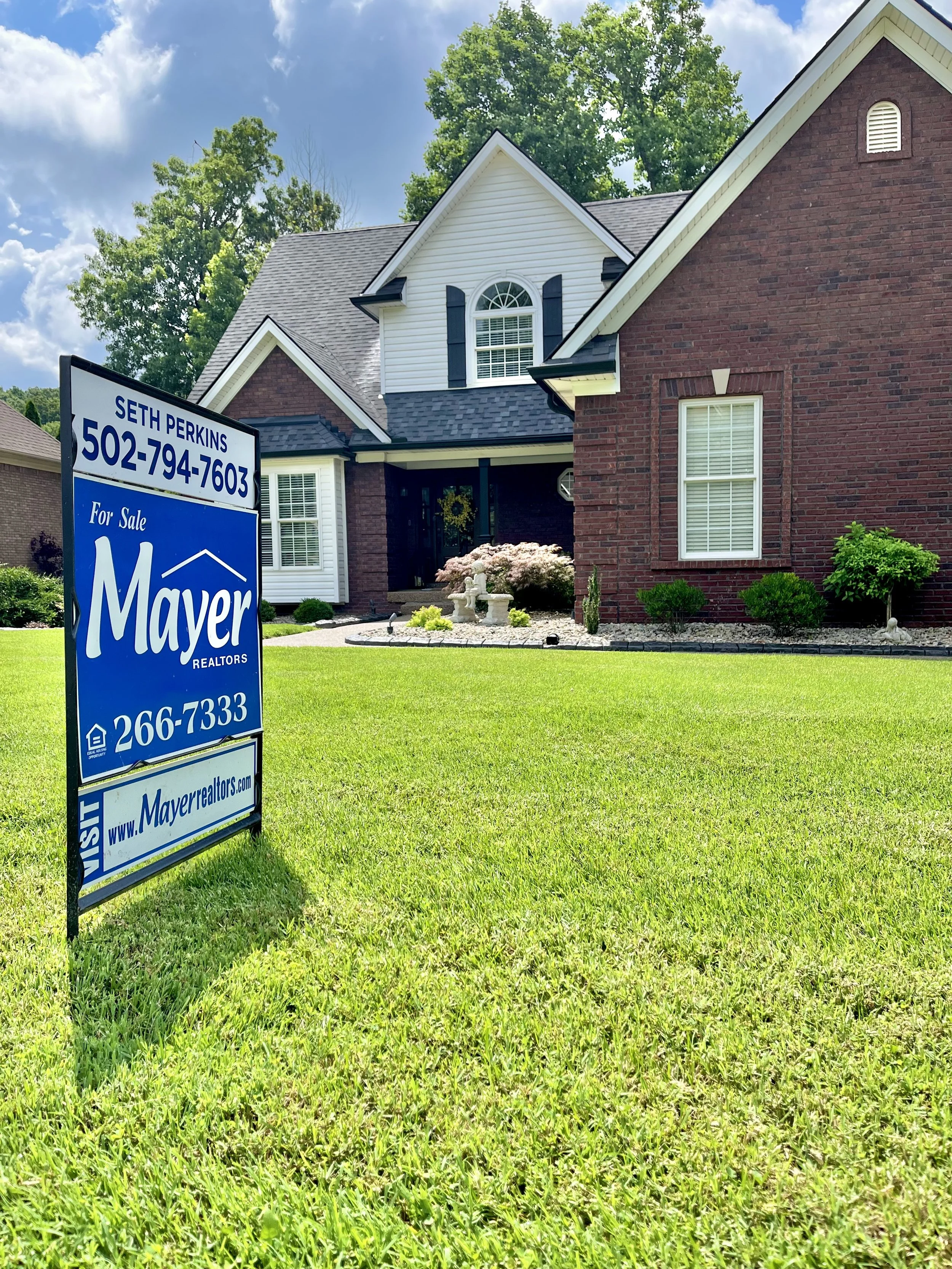 A house with a mix of brick and white siding, with green landscaping and a for-sale sign in the yard.