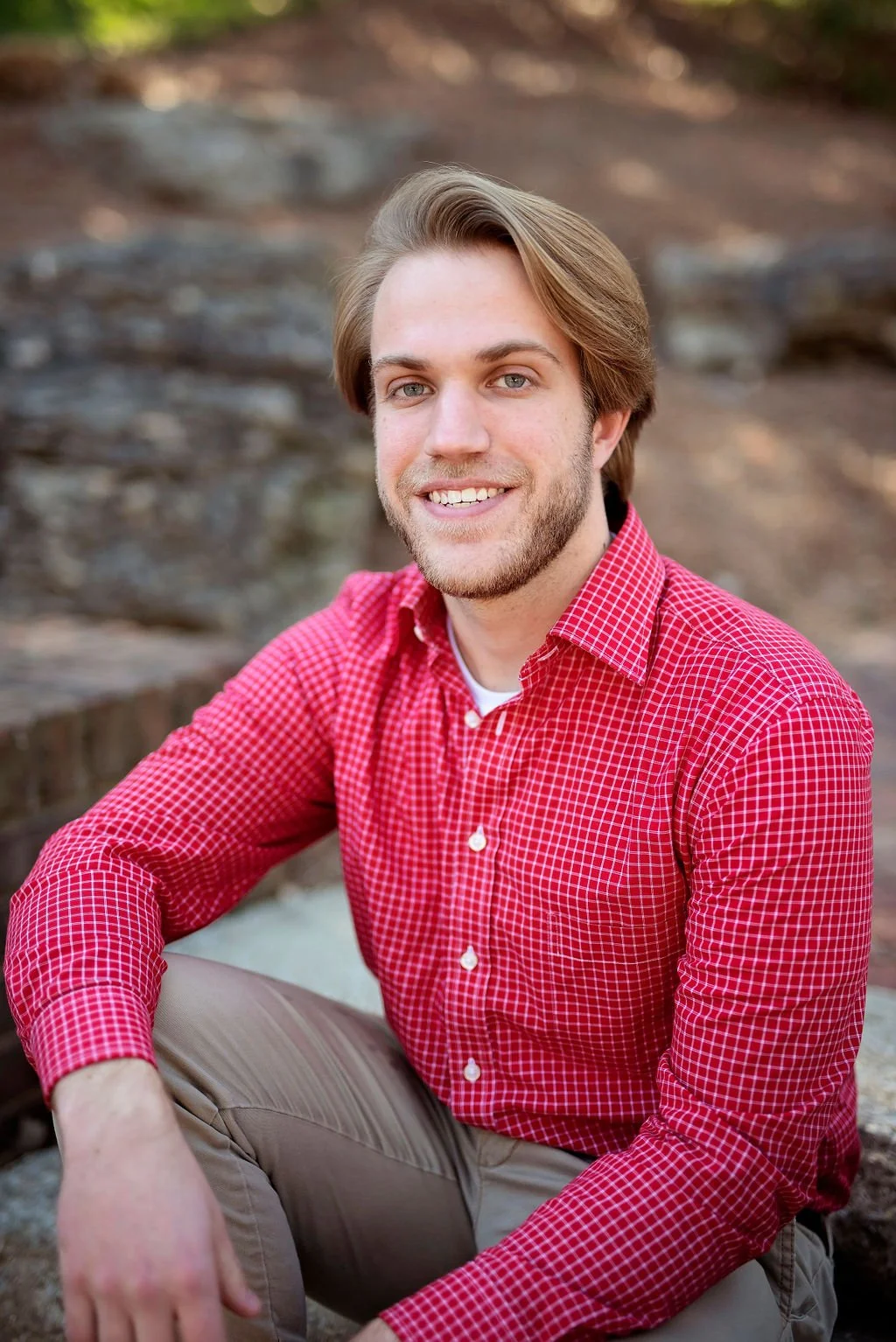 Portrait of a young man with light brown hair, blue eyes, and a light beard, smiling and wearing a red checkered shirt, sitting outdoors with a blurred natural background.