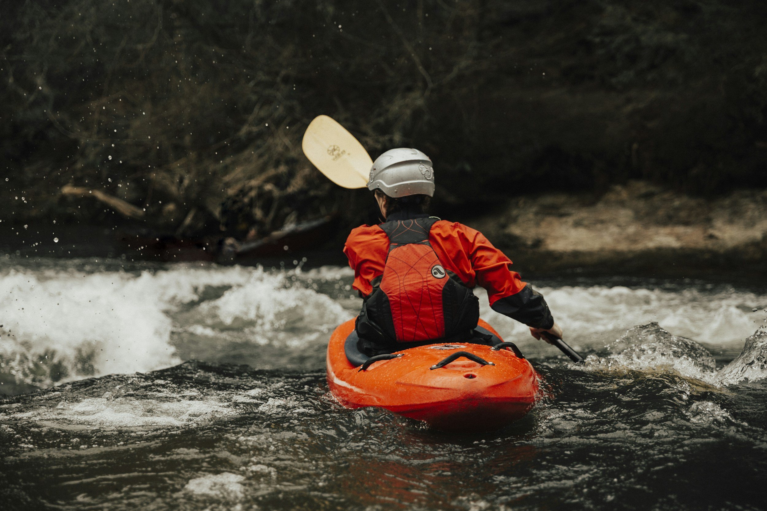 Person kayaking on white water river with rocky banks, wearing a helmet and orange life jacket, paddling downstream.