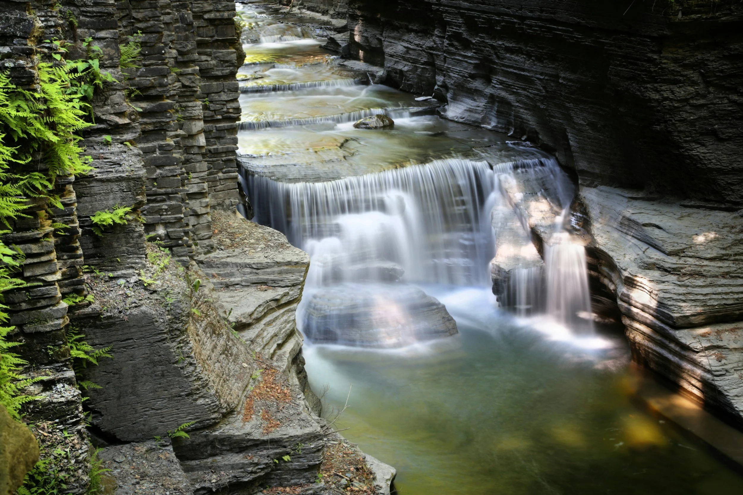 A small waterfall flowing over layered rocks in a narrow canyon with green ferns on the left side.