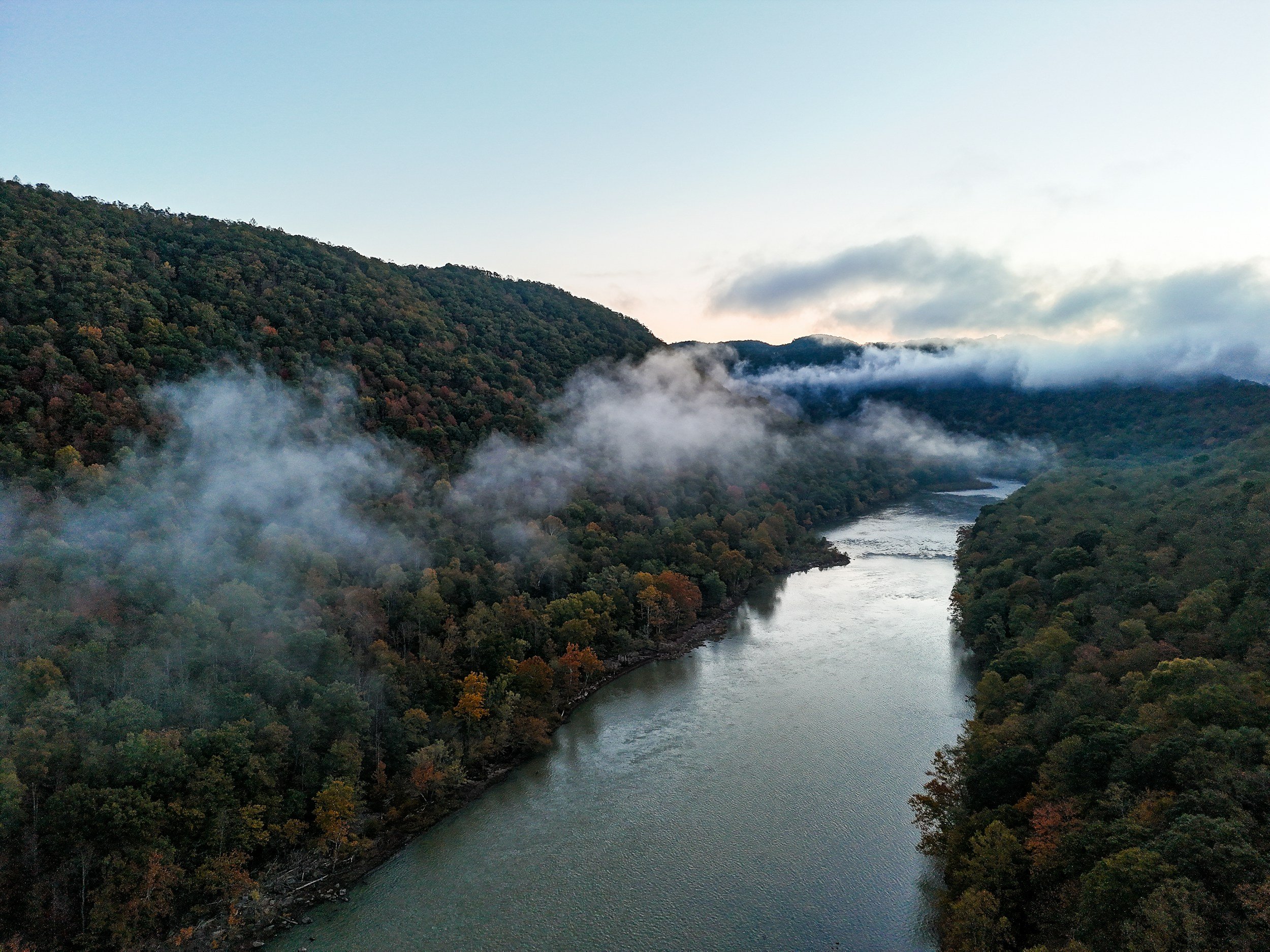A river flowing through a valley with hills on each side covered in trees, some trees showing autumn colors, with mist or fog rising from the river and hills under a clear sky.