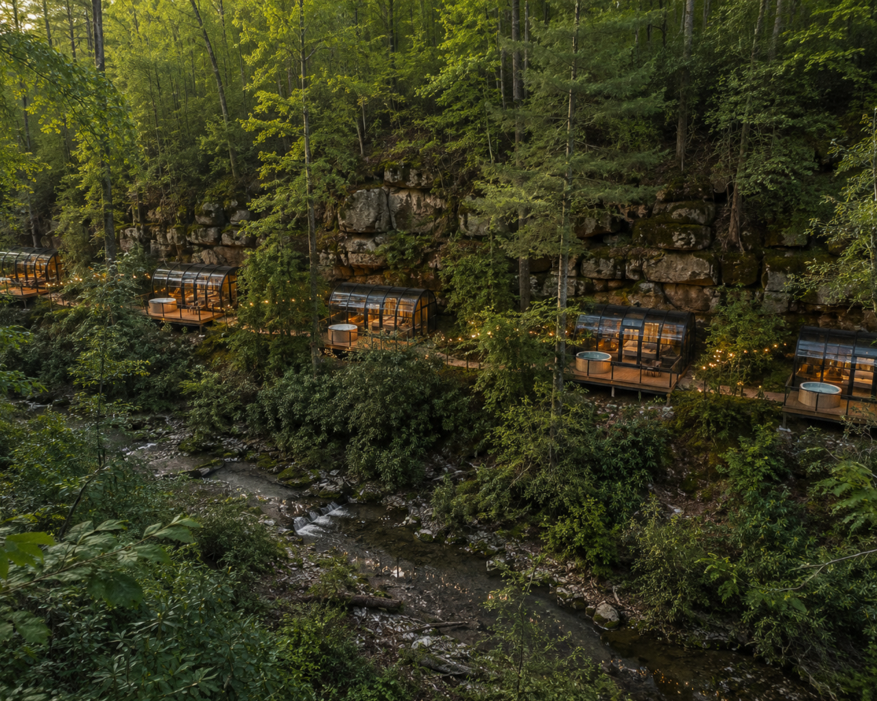 Series of glass-enclosed outdoor pods with seating along a wooded hillside at dusk, overlooking a small river.