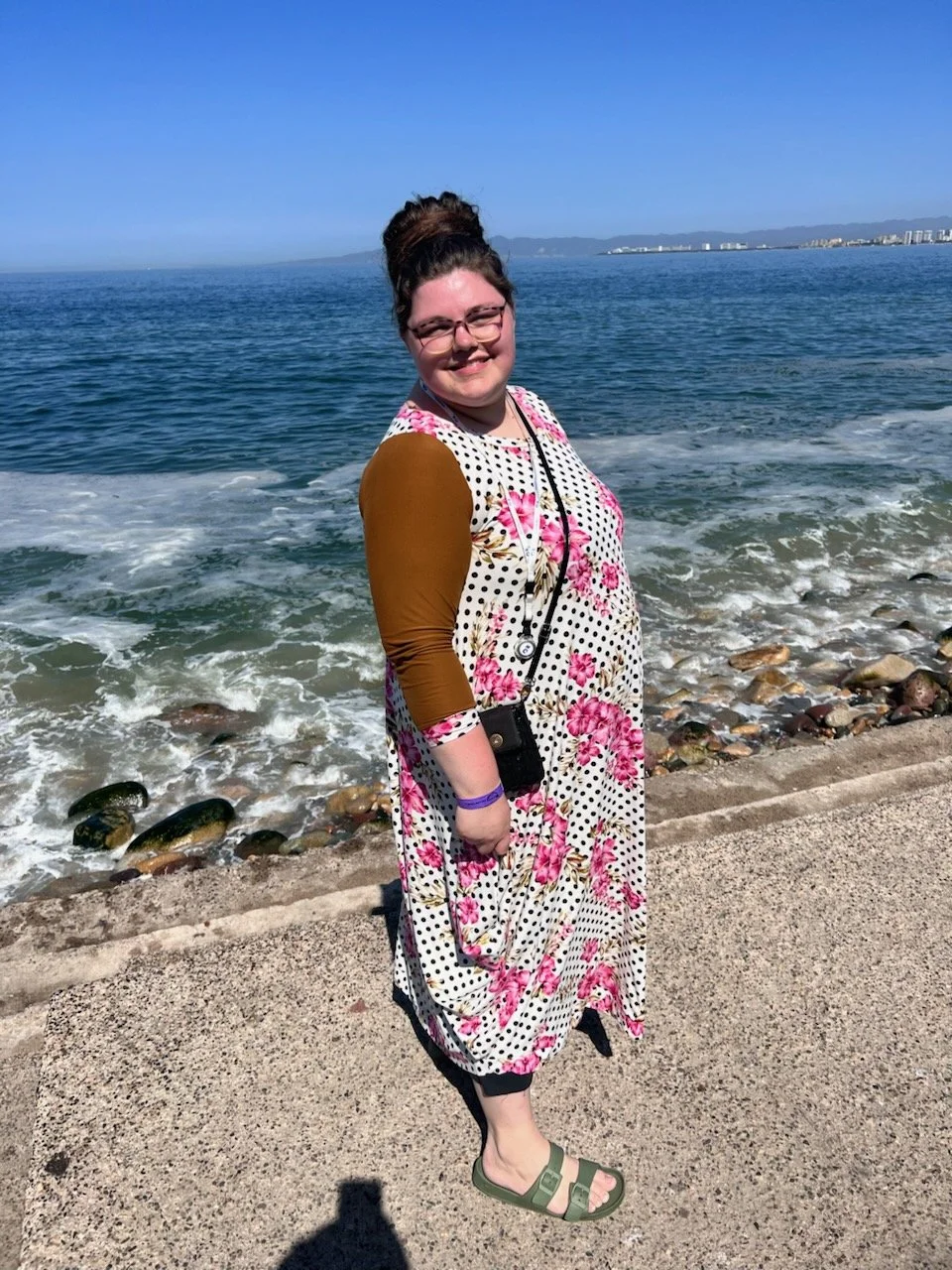 A woman standing on a concrete path by the ocean, smiling, with mountains and cityscape in the background on a sunny day.