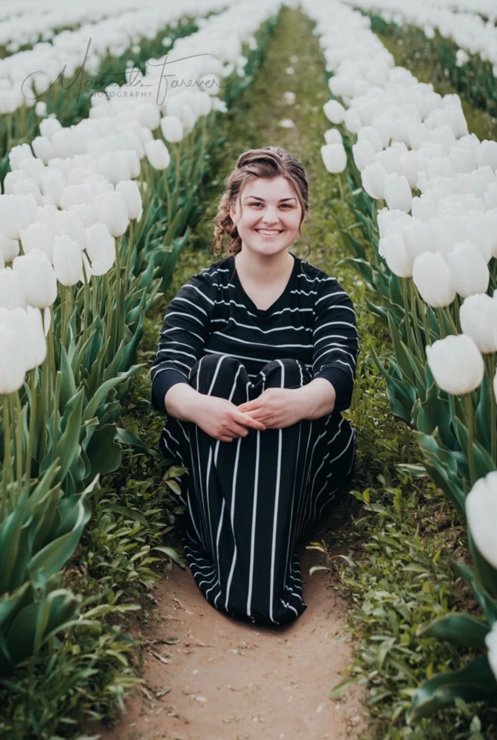 A smiling woman in black and white striped clothing sitting in the middle of a path surrounded by white tulips in a garden.