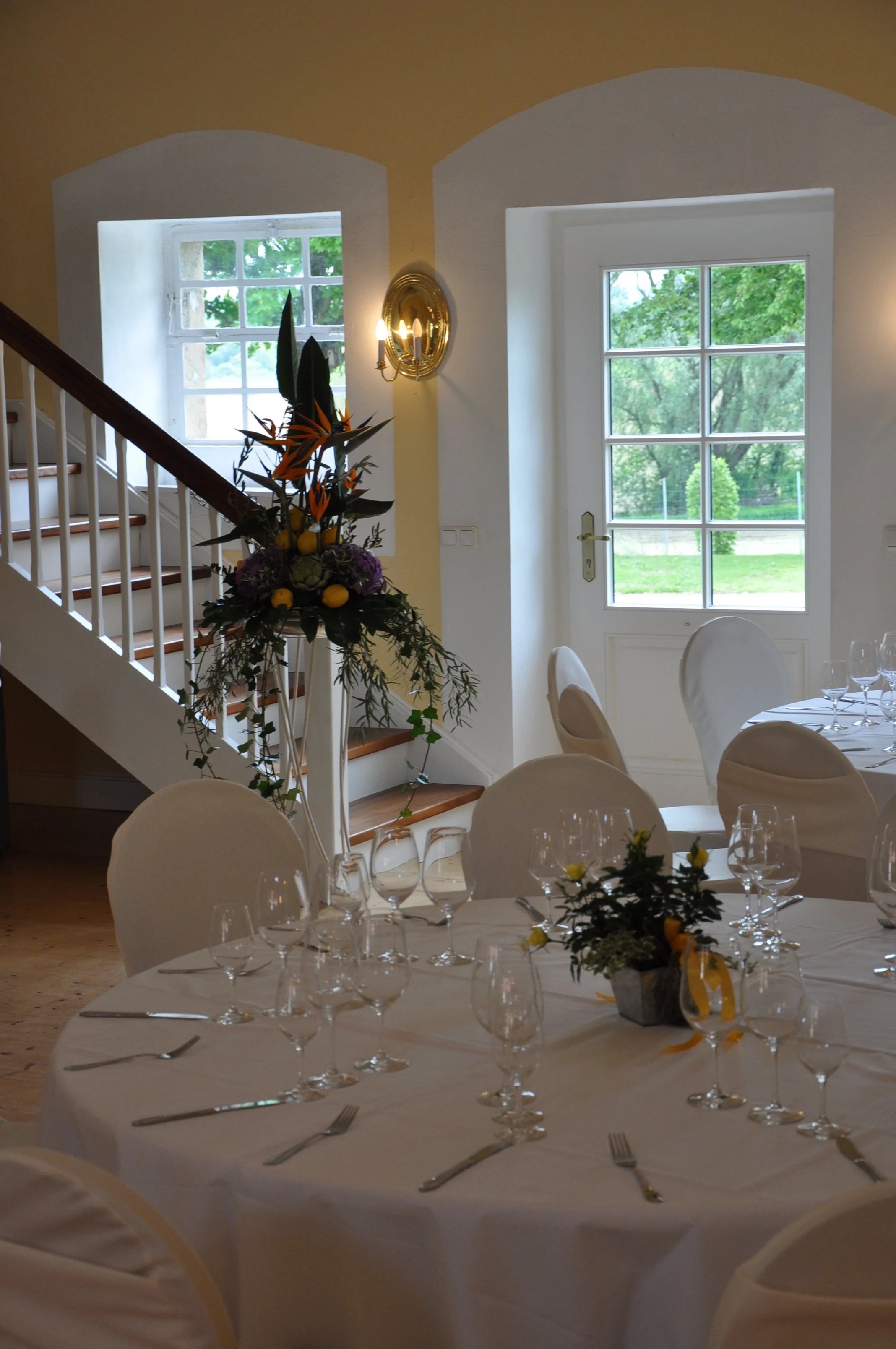 Ein festlich gedeckter Tisch mit weißen Tischdecken und mehreren Weingläsern, in einem Raum mit großen Fenstern und einer Dekoration mit Blumen und Kerzen, wahrscheinlich für eine Feier oder ein Event.