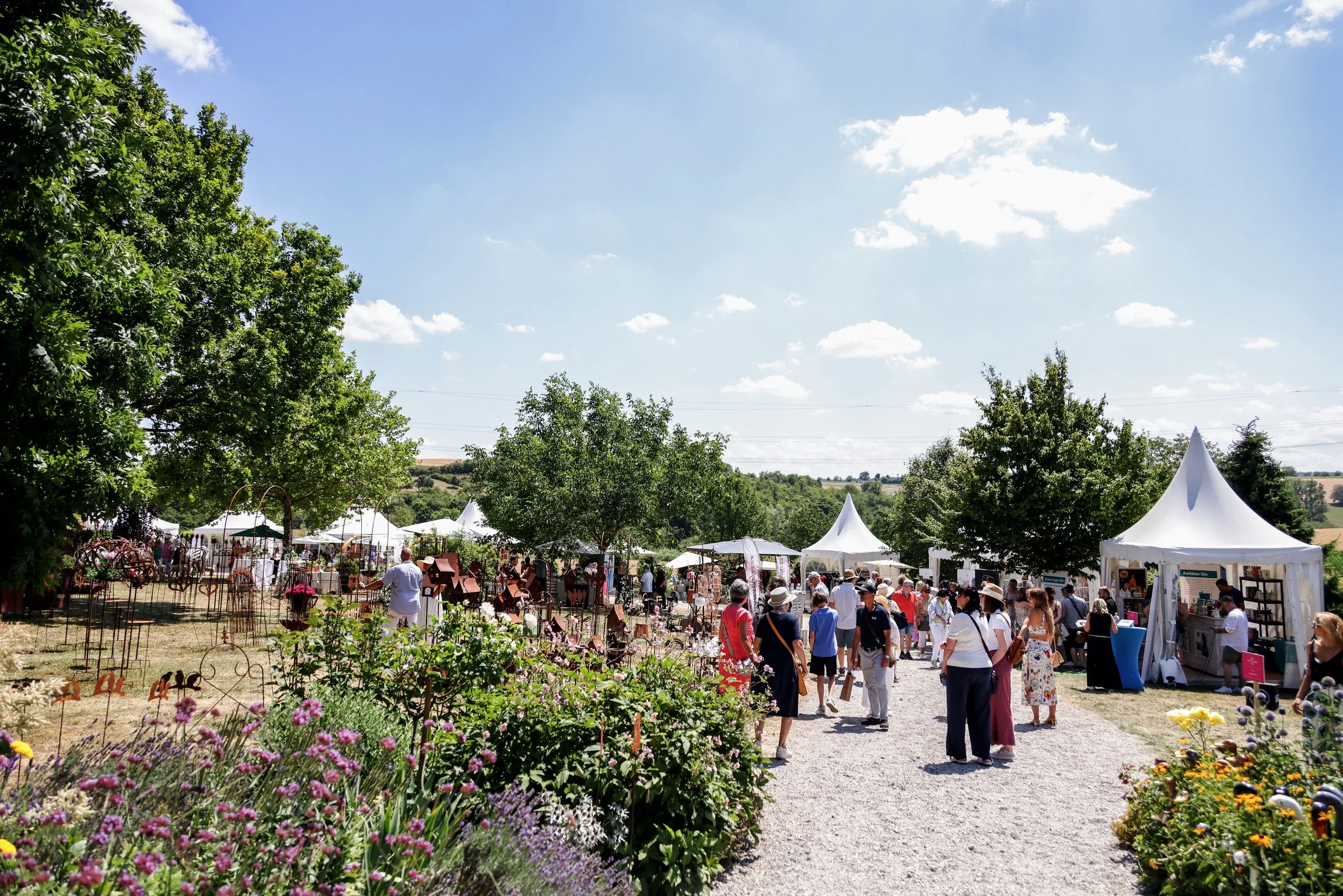 Ein Fest im Freien mit weißen Zelten, vielen Menschen und buntem Blumenbeet im Vordergrund, bei sonnigem Himmel.