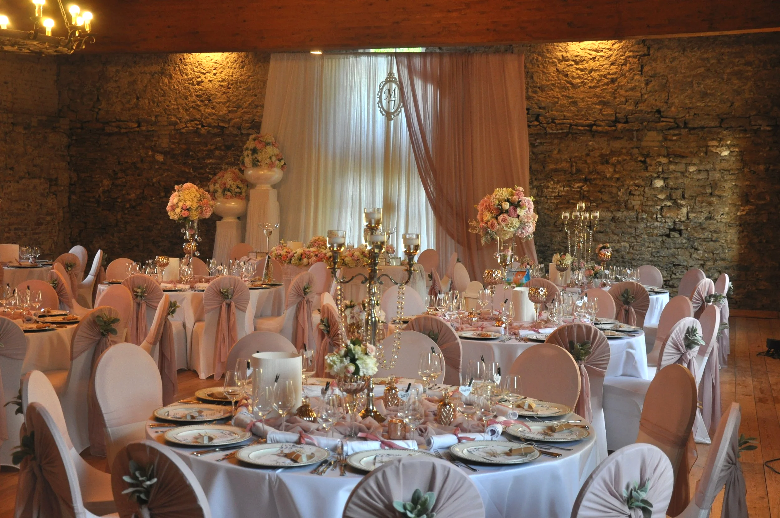 Elegant geschmückter Festsaal mit runden Tischen, Blumensträußen und Kerzen, für eine besondere Veranstaltung wie eine Hochzeit, mit einer Kombination aus warmem Licht, einem steinernen Wand und weichen Stoffvorhängen.