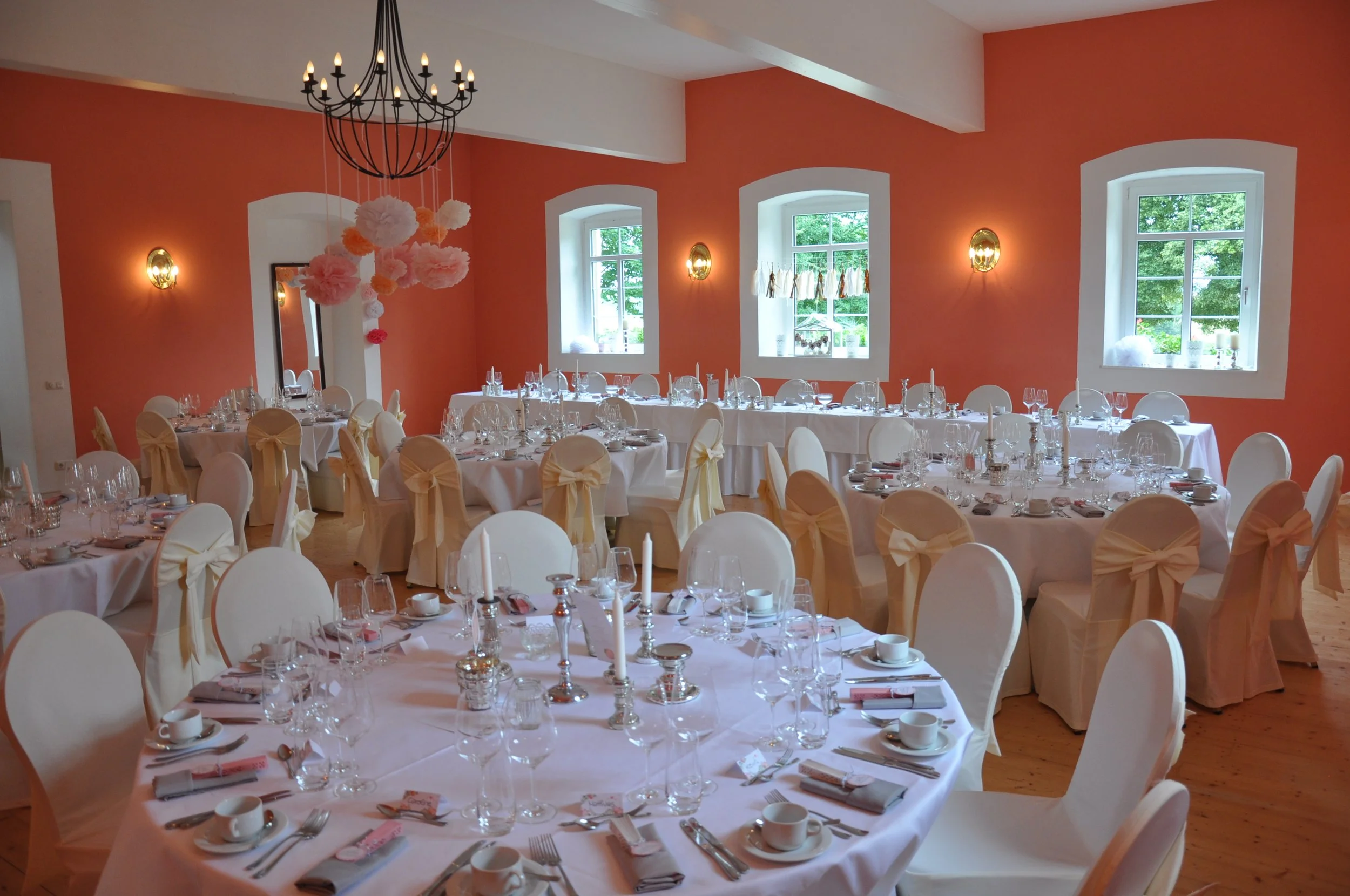 Festlich gedeckter Saal mit runden und rechteckigen Tischen, weißen Tischdecken, Kerzen, Gläsern, Geschirr und Besteck, goldenen Scheinwerfern an der Wand, großen Fenstern mit Blick auf grüne Bäume, einer schwarzen Kronleuchterdecke, rosa Dekoratione