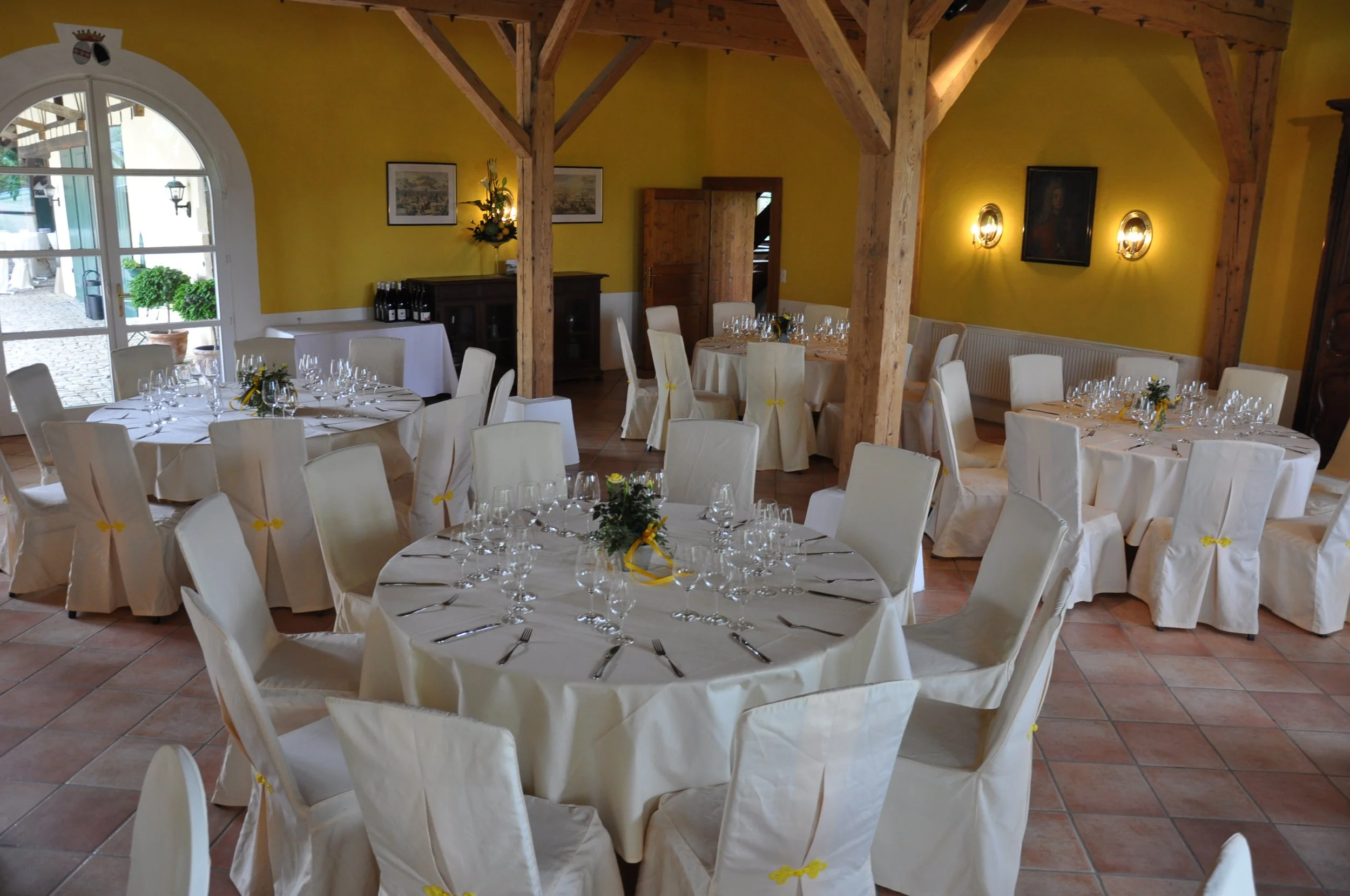 Ein festlich für ein Essen gedeckter Raum mit weißen Tischtüchern, Weingläsern und kleinen Blumenarrangements auf Tischen, gelbe Wände, Holzbalken und Tageslichteinfall durch große Fenster.