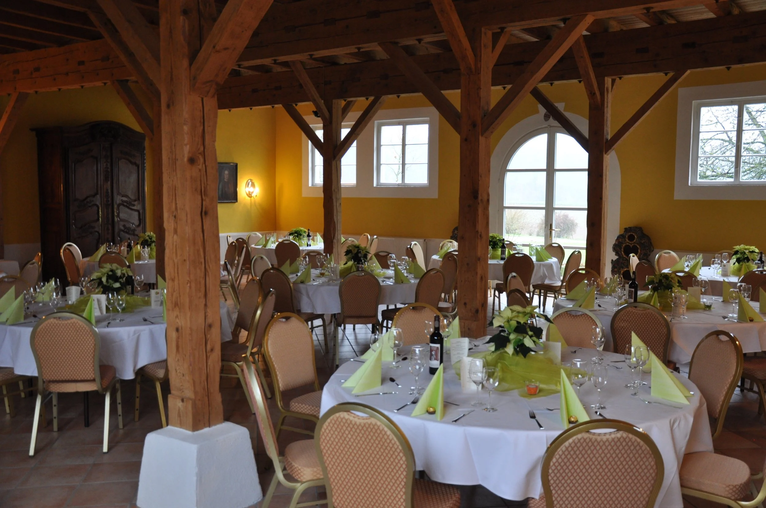 Ein festlich gedeckter gemeinsamer Tische im Saal eines Restaurants. Es sind mehrere runde Tische mit weißen Tafeldecken, Kerzen, Gläser, Besteck und grüne Servietten. Im Hintergrund sind große Fenster, die viel Tageslicht in den Raum lassen, und Hol