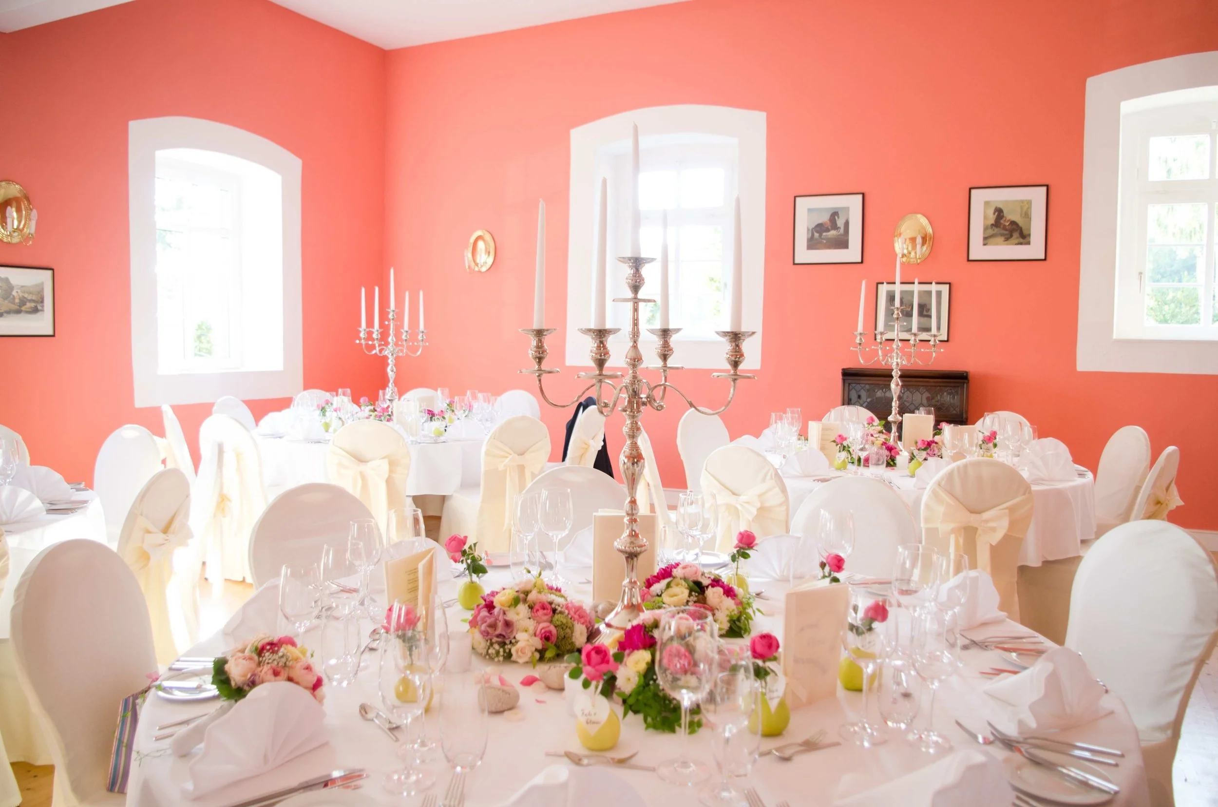 Ein festlich gedeckter Raum mit runden Tischen, weißen Stühlen, Kerzenleuchtern und bunten Blumenarrangements. Die Wände sind rosafarben mit Bildern und Fenstern, die Tageslicht hereinlassen.