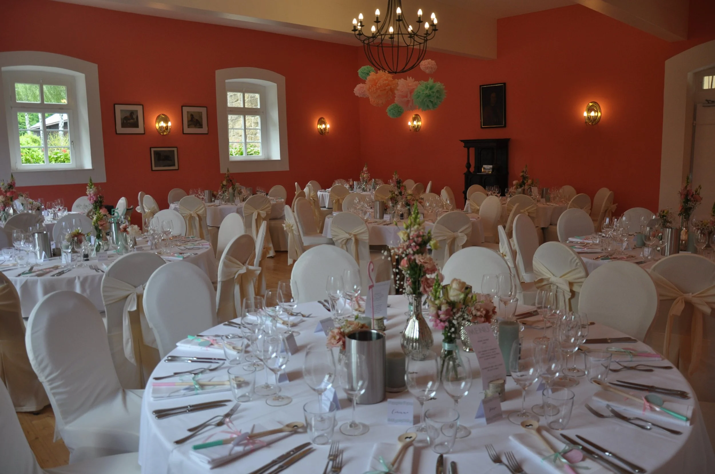 Der Saal ist festlich gedeckt für eine Feier, mit weißen Tischen und Stühlen, Blumenarrangements, Wein- und Wassergläsern, Besteck und Menükarten. Es gibt Fenster, die Tageslicht hereinlassen, und die Wände sind in einem warmen Rot gehalten. Eine gro