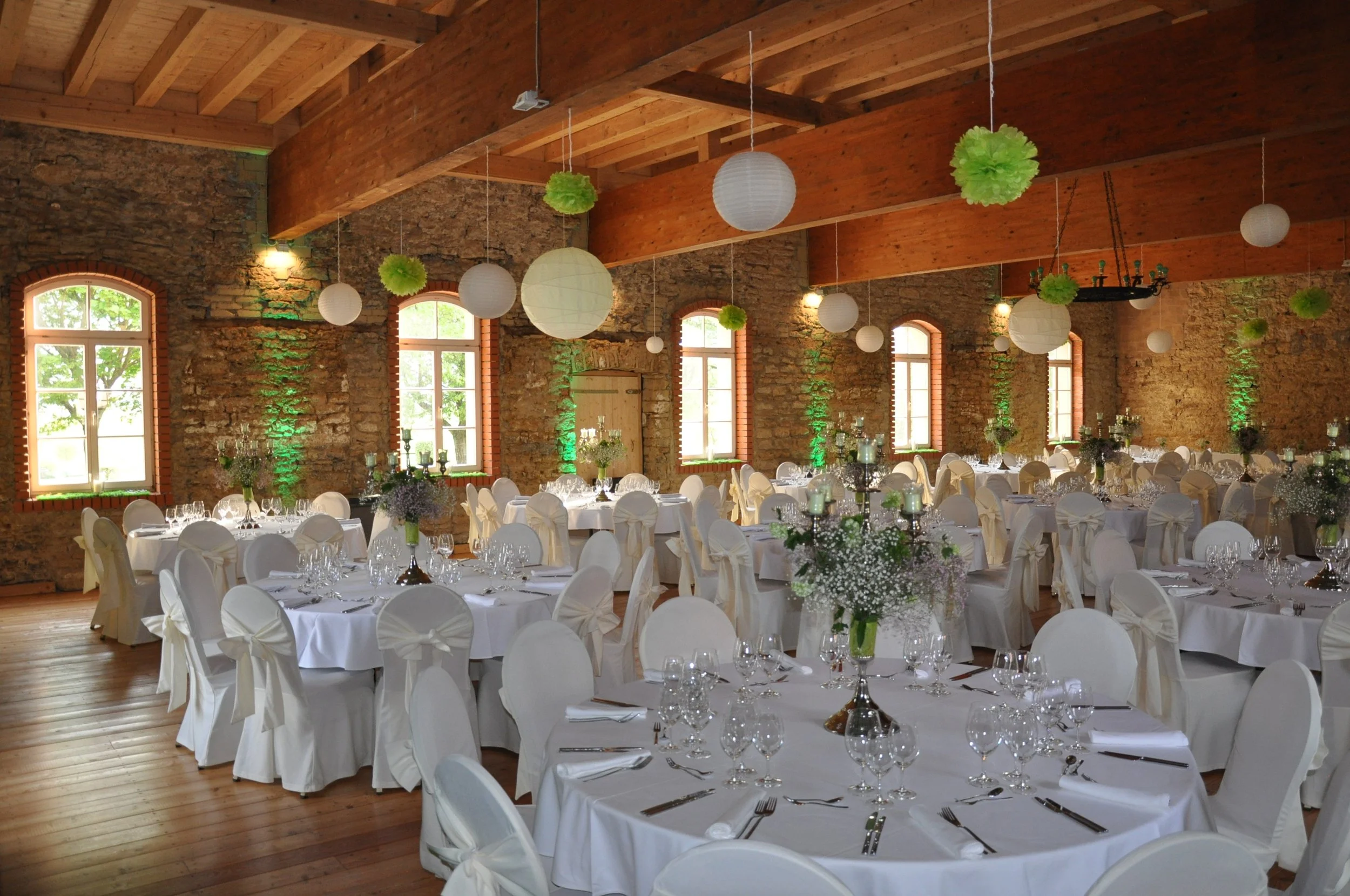 Ein festlich gedeckter Saal mit runden Tischen, weißen Stühlen und Blumenarrangements, große Fenster mit natürliche Beleuchtung, Holzdecke und Backsteinwände, Dekoration mit grünem Licht