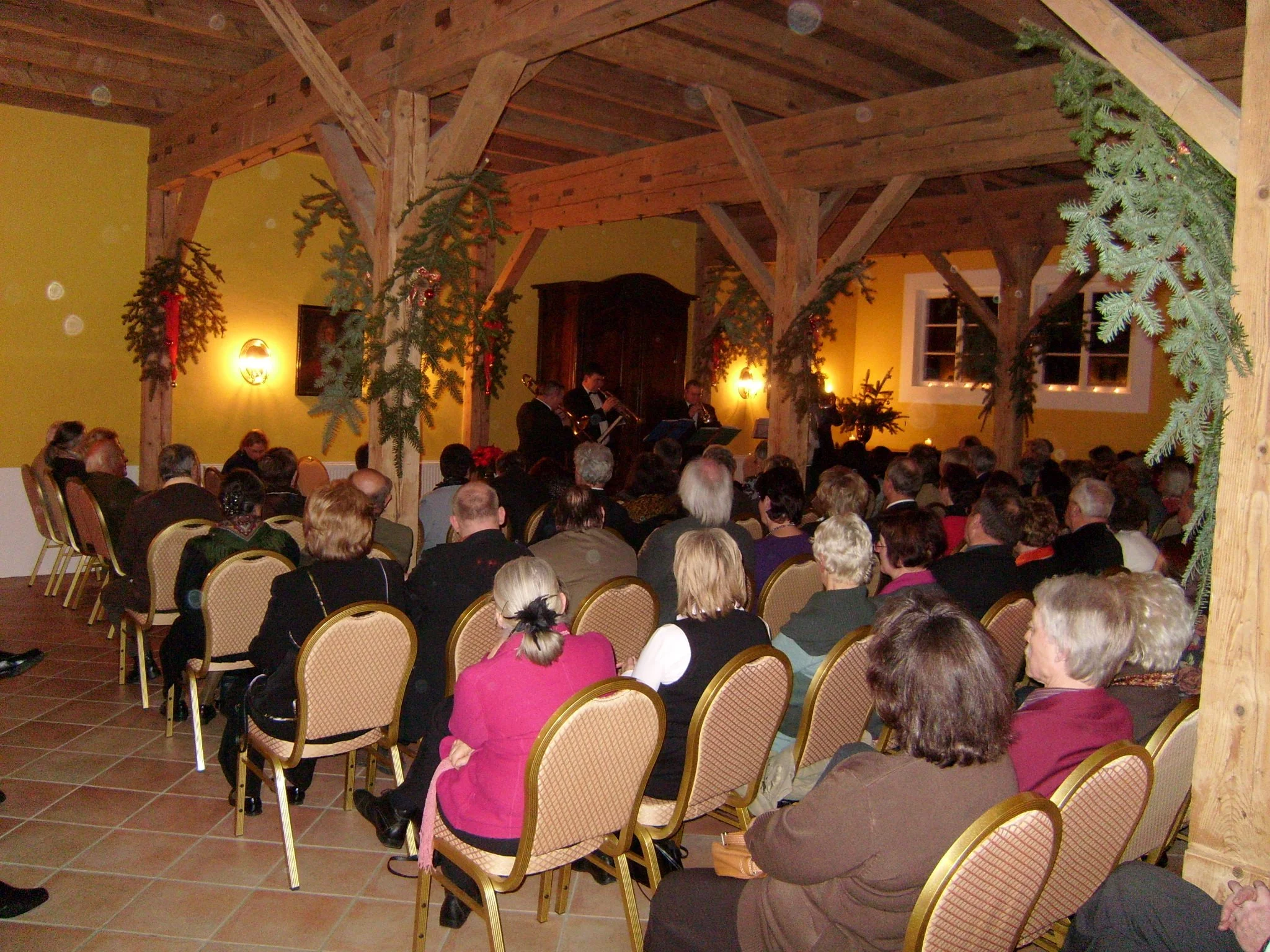 Ein festlich dekorierter Veranstaltungsraum mit Anwesenden, die einem musikalischen Konzert zuhören. Der Raum hat eine holzgetäfelte Decke, gelbe Wände und ist mit Tannenbaumzweigen und Kerzen geschmückt.