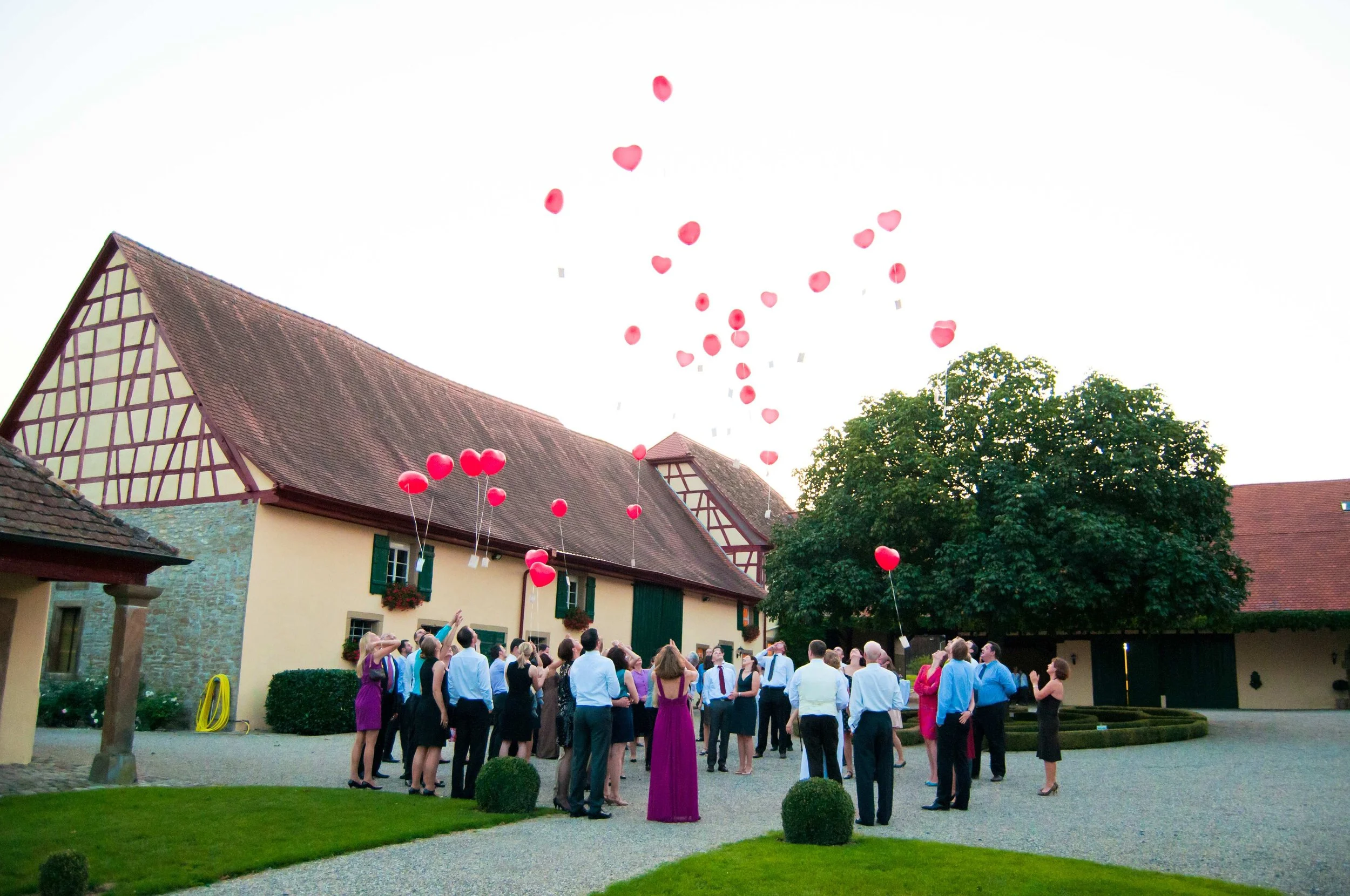 Eine Gruppe von Menschen in formeller Kleidung sieht während eines Abends in einem Hof Gebiet zu, während Heliumballons in Herzform in den Himmel steigen, vermutlich bei einer Feier oder Hochzeit.