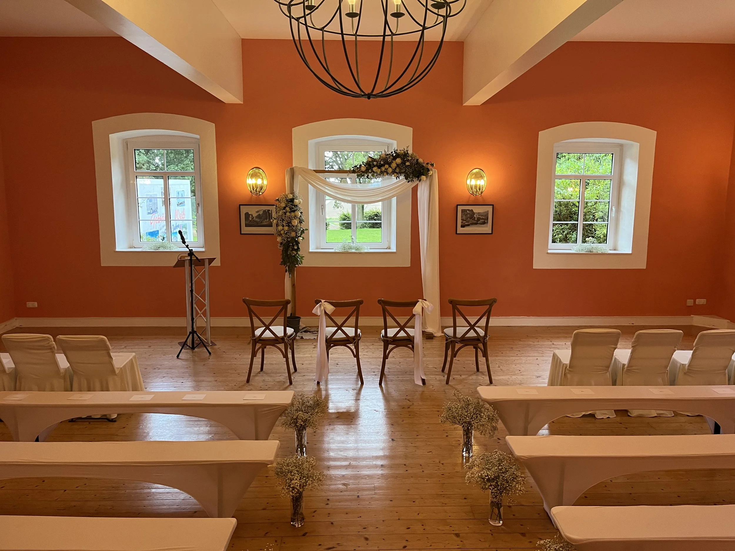 Der Raum ist für eine Hochzeitszeremonie vorbereitet, mit weißen Stühlen, einem Altar mit weißen Tüchern und Blumen, drei hohen Fenstern mit Blick ins Grüne, einem Kronleuchter an der Decke und warmen Wandlampen.