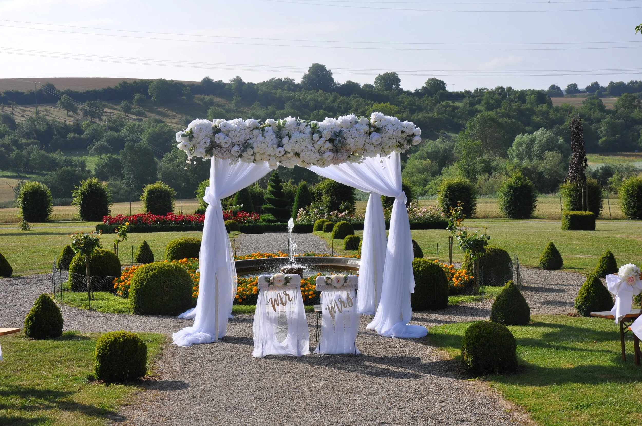 Ein Hochzeitshintergrund im Freien mit weißen Blumen und weißen Vorhängen, umrahmt von Büschen, bei sonnigem Wetter in einer ländlichen Umgebung.