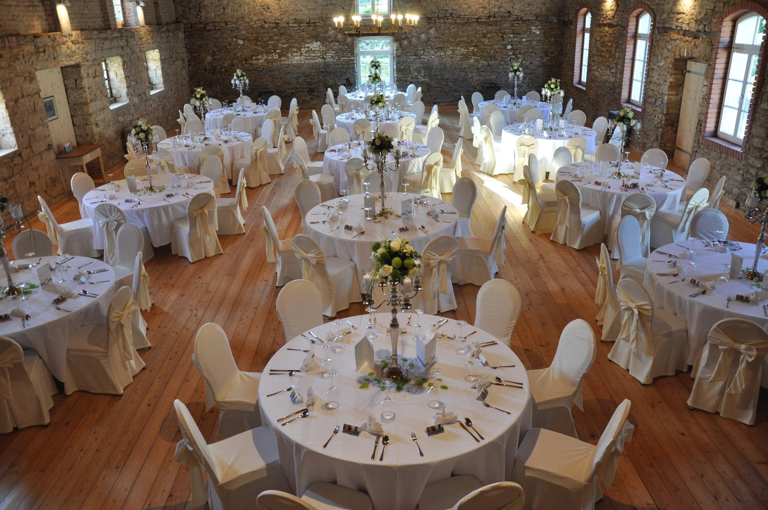 Ein festlich gedeckter Saal mit runden Tischen, weißen Stuhlhussen und eleganten Kerzen- und Blumenarrangements, eingerichtet für eine Feier oder Hochzeit in einem Raum mit Ziegelmauern und Holzboden.