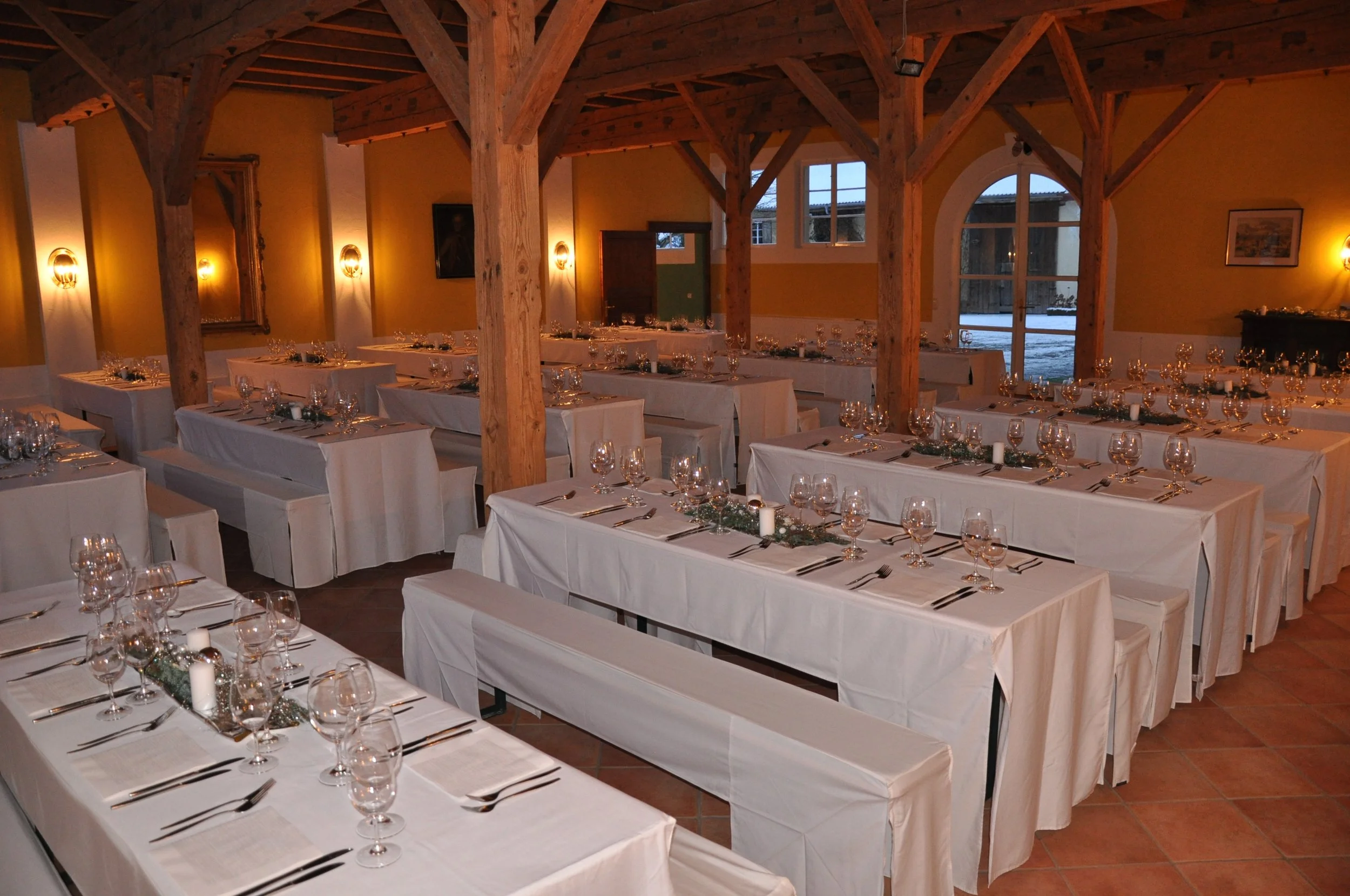 Ein festlich gedeckter Speisesaal mit weißen Tischtüchern, zahlreichen Weingläsern, Kerzen und silbernem Dekor, in einem Raum mit Holzdecke und großen Fenstern, draußen liegt Schnee.
