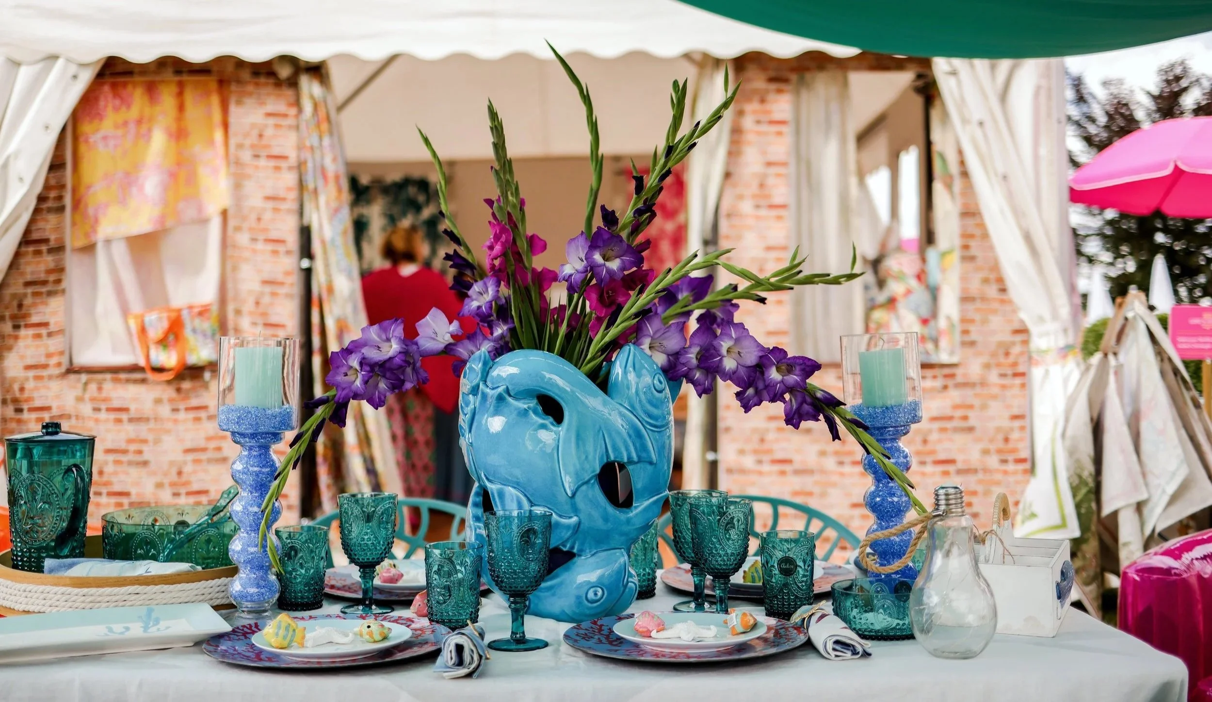 Pagodenzelt der Ausstellerin Solenzara bei der LIFE'S FINEST; Bunter festlicher Tisch mit blauer Vase und violetten und lila Blumen, grünen Gläsern, Kerzen, Geschirr und Dekorationen im Freien, mit einem Zelt und einer Brickwand im Hintergrund