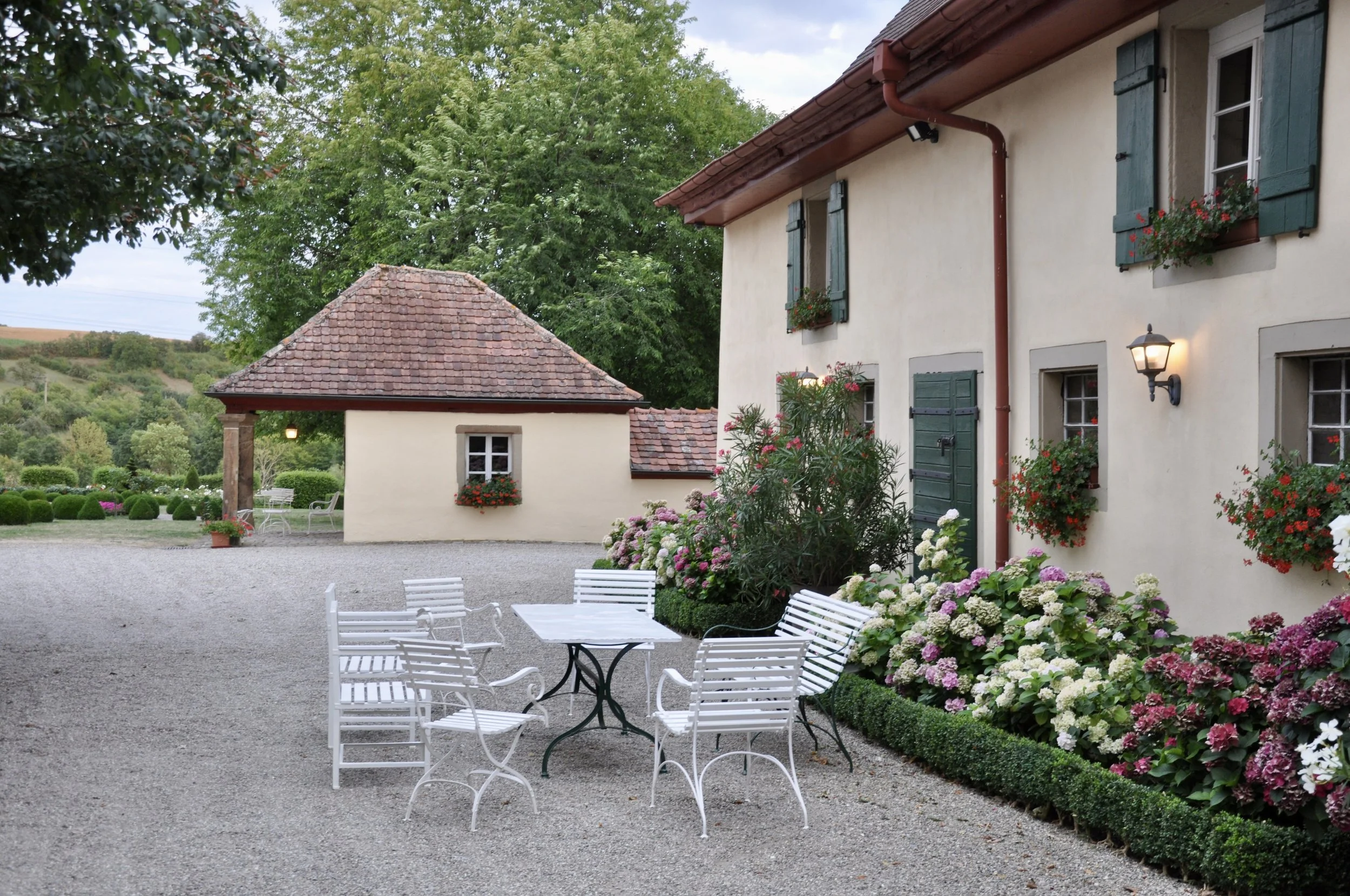 Ein ländliches Haus mit einer Terrasse, umgeben von bunten Blumen und grünen Bäumen, bei Sonnenuntergang.